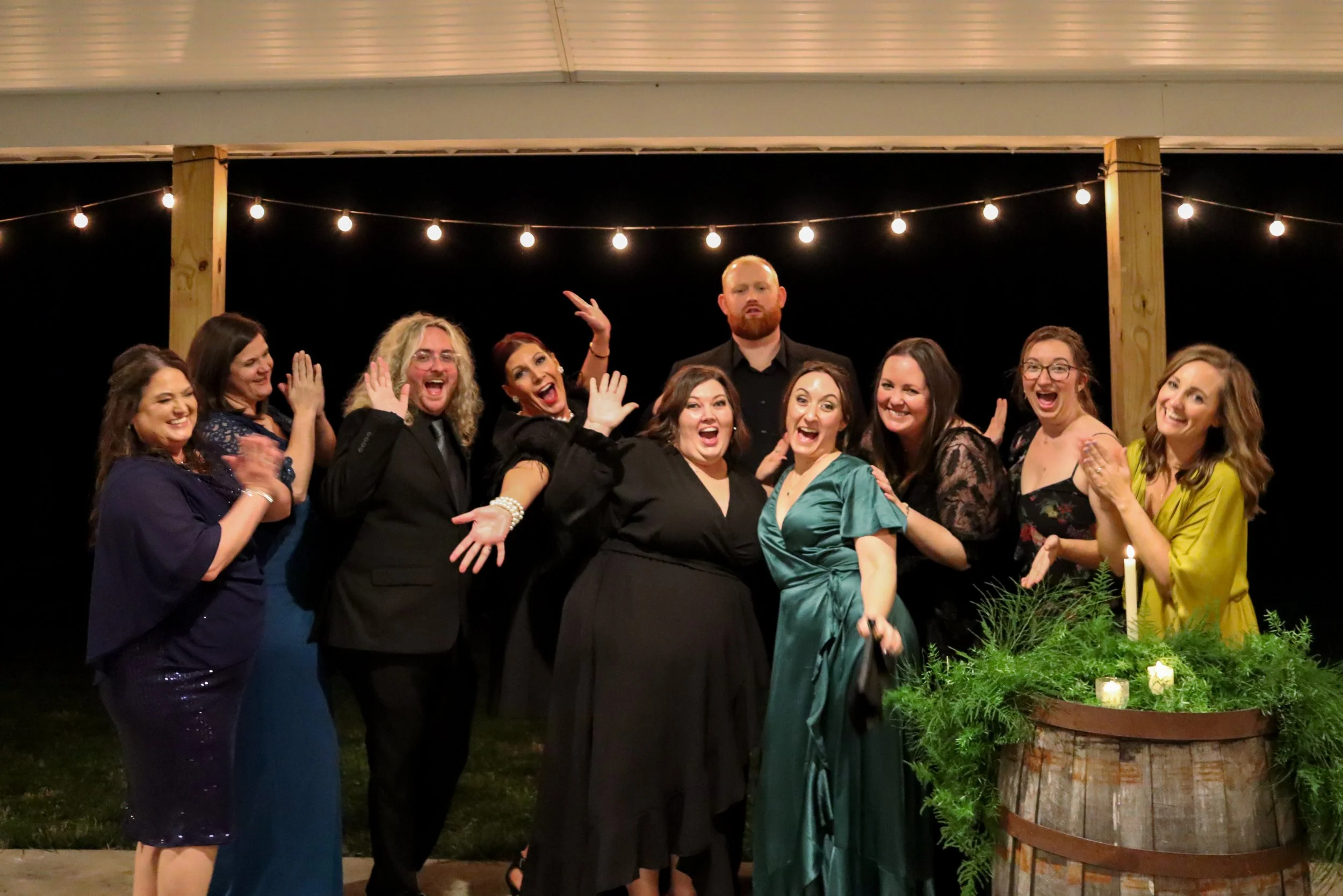 A group of ten people, mostly women, celebrating at night under string lights. They are dressed in formal and semi-formal attire, smiling and posing happily. There are candles and greenery in the foreground.