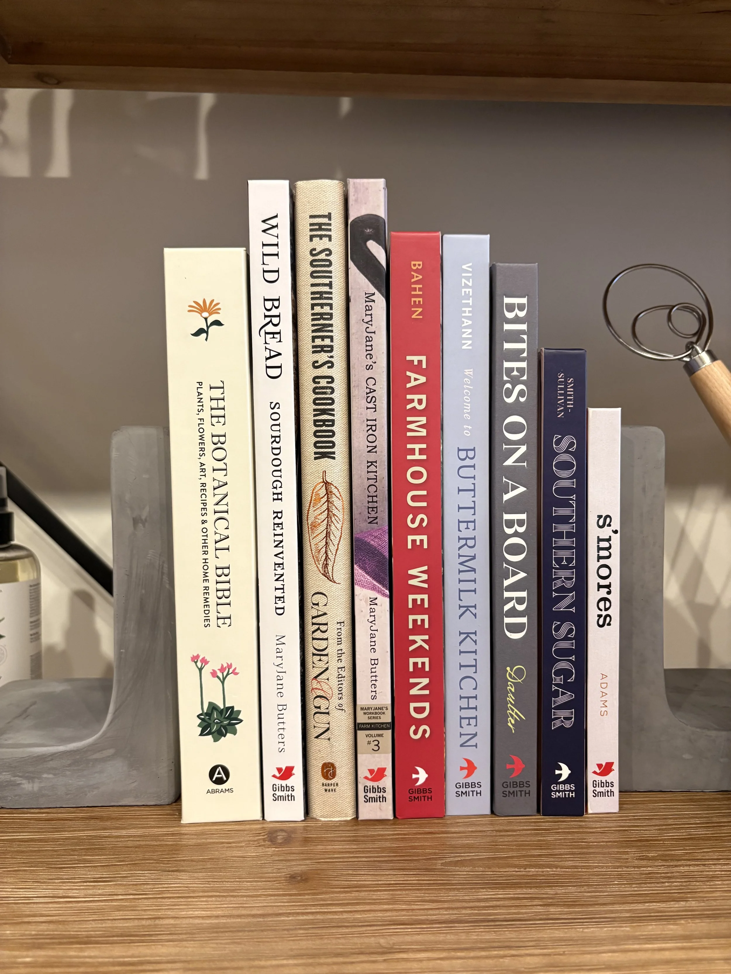 A row of books on a wooden shelf with titles including The Botanical Bible, Wild Bread, The Southern Farmer's Cookbook, MarryJane’s Cast Iron Kitchen, Farm Kitchen, Bake in the War, Vise Than to Butcher, The Way to Beekeepers, Kids in the Kitchen, Butter, Soup and Sulaiman’s Sugar.