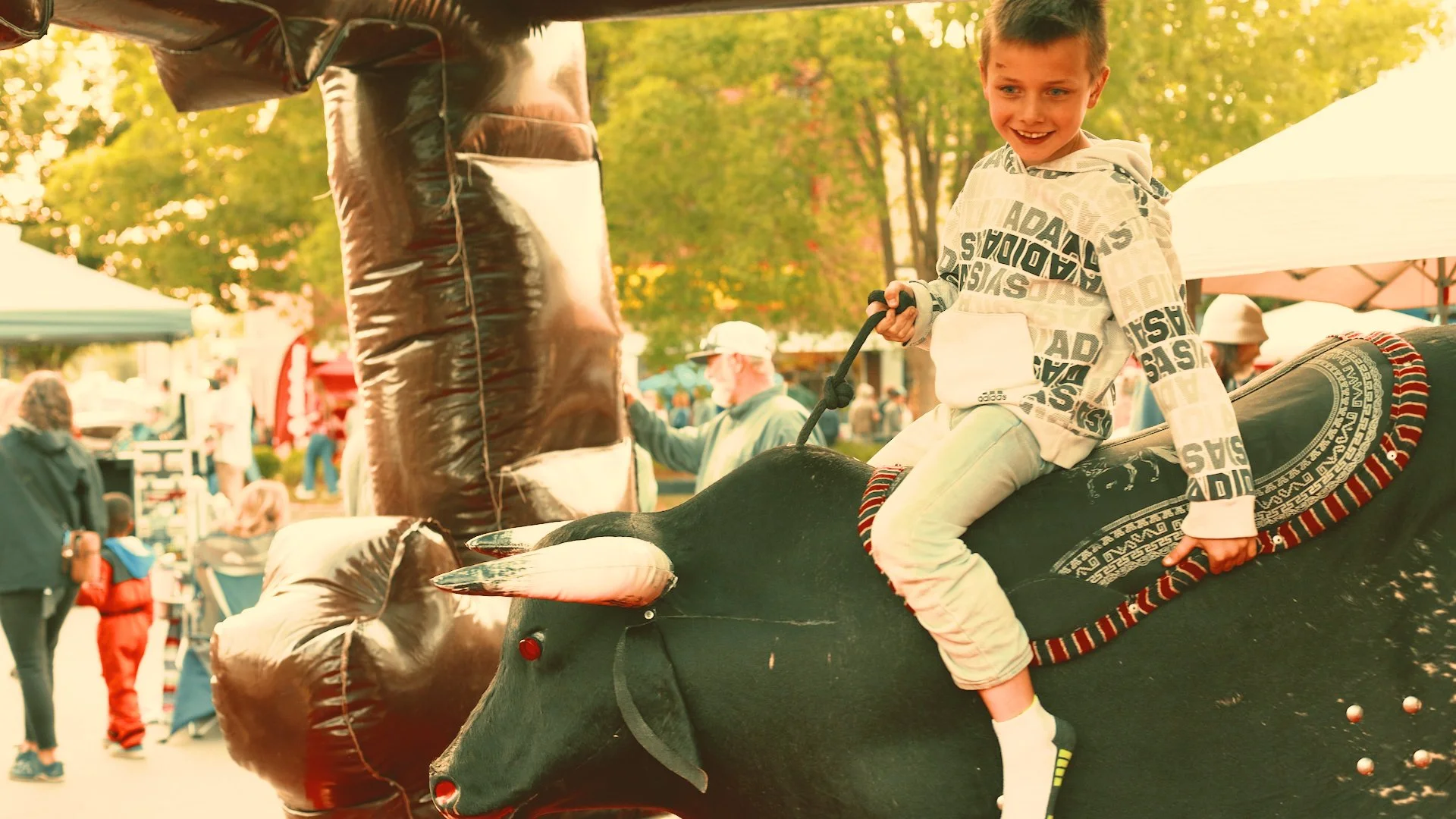 A young boy with short brown hair and a white hoodie with black text is sitting on a large, black inflatable bull at an outdoor event or fair during sunset. The background shows people, trees with green leaves, and tents or booths.