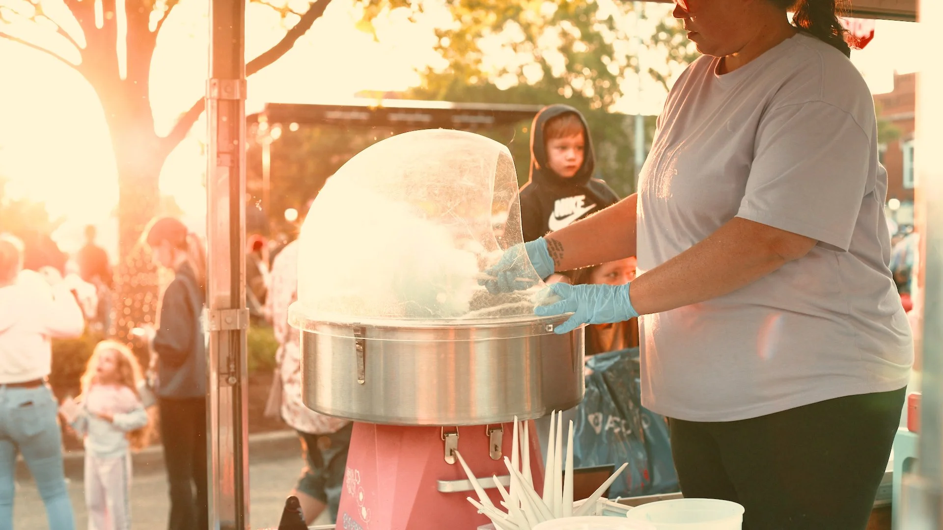 Person making cotton candy at an outdoor fair during sunset with children and other people in the background.