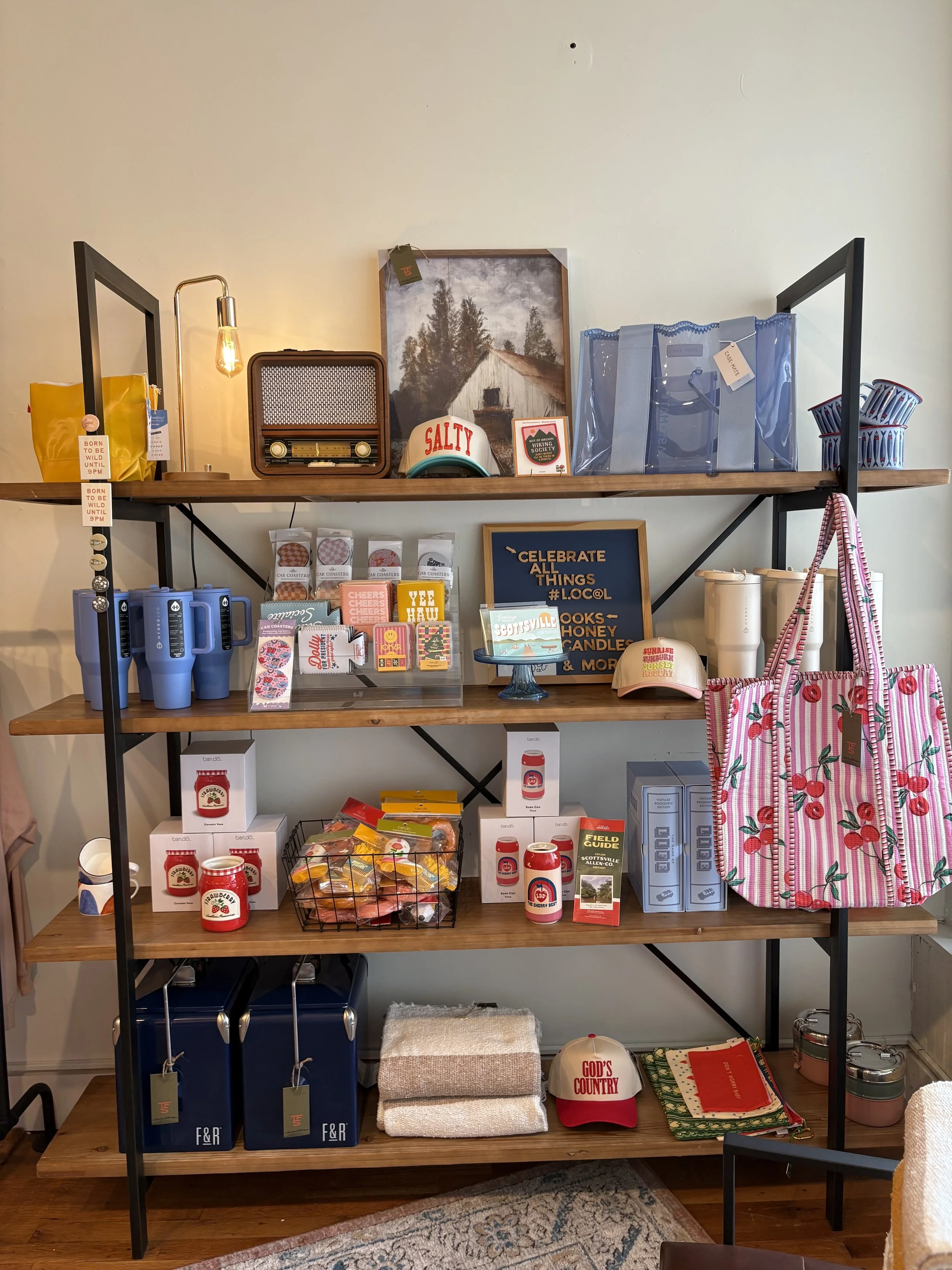 A black metal and wood shelving unit with four shelves displaying various decorative and practical items, including a hat, framed artwork, books, kitchenware, and small accessories, in a cozy interior setting.