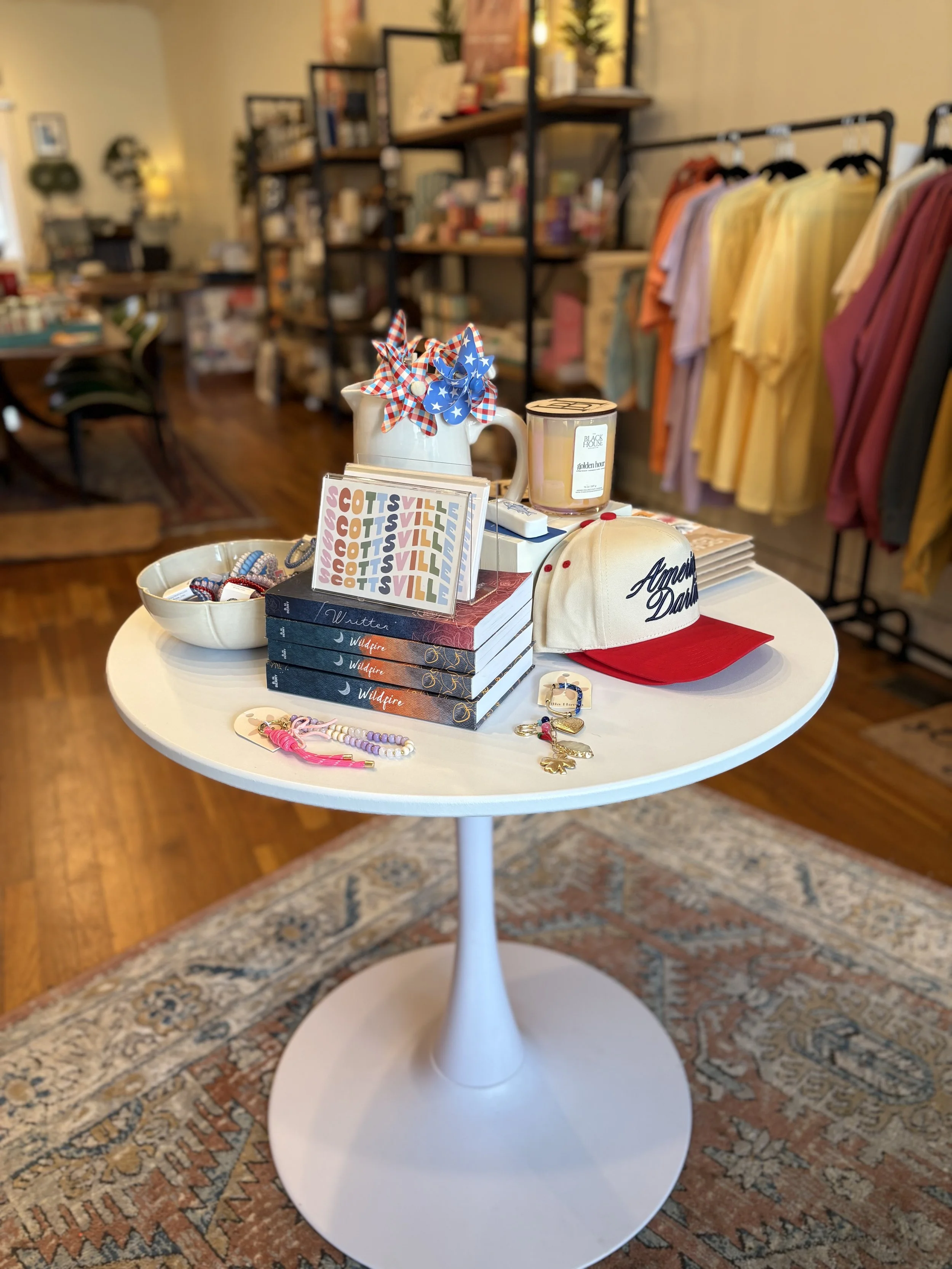 A round white table displaying various accessories including bracelets, a baseball cap, and a small dish with keychains. In the background, there are shelves with books, decorative items, and a clothing rack with colored shirts. The room has wooden flooring and a patterned rug under the table.