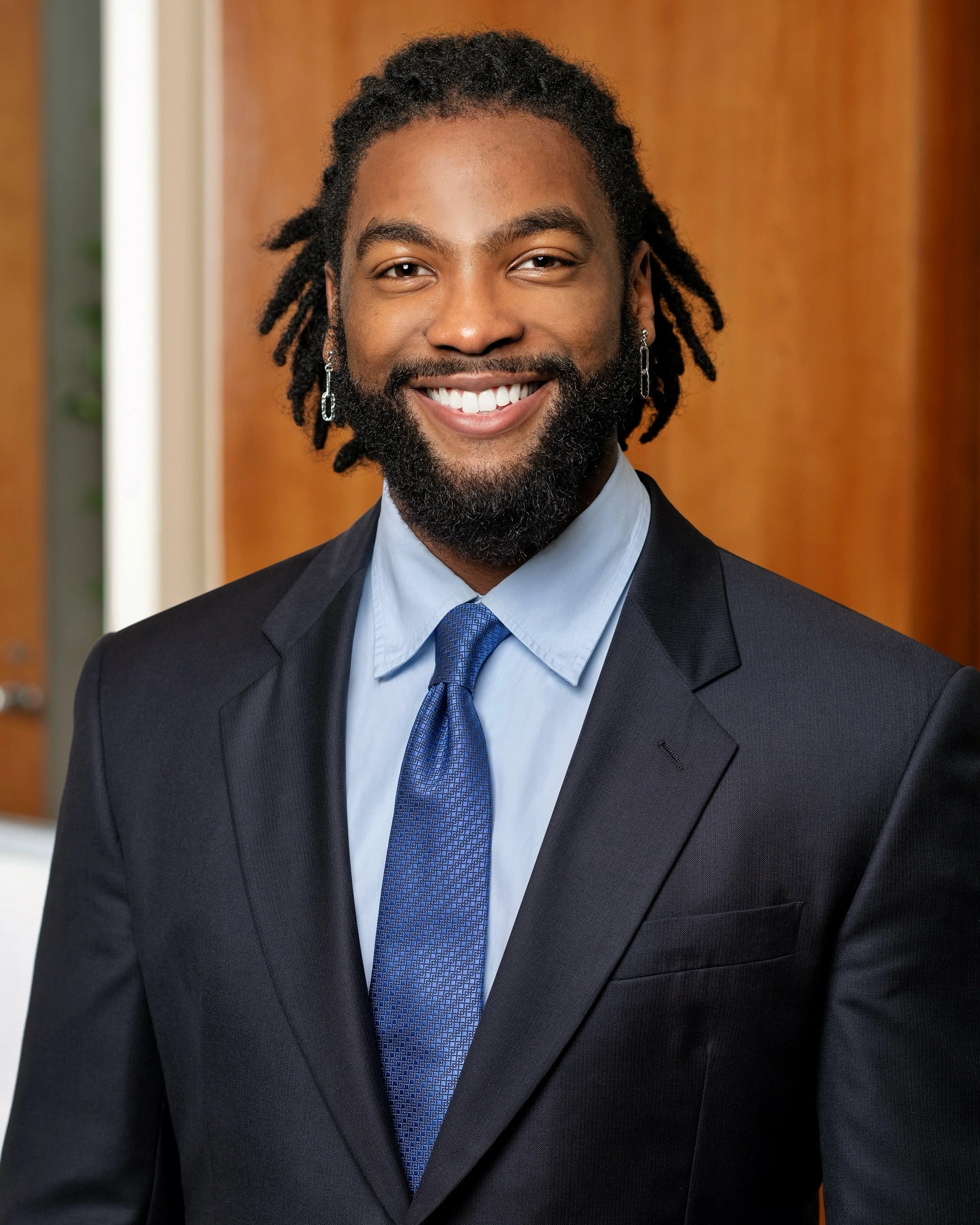 A man dressed in a dark suit, light blue shirt, blue tie, with curly hair and earrings, smiling, in front of a wooden background.