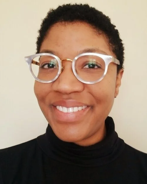 A woman with short curly hair wearing clear glasses and a black turtleneck, smiling at the camera.