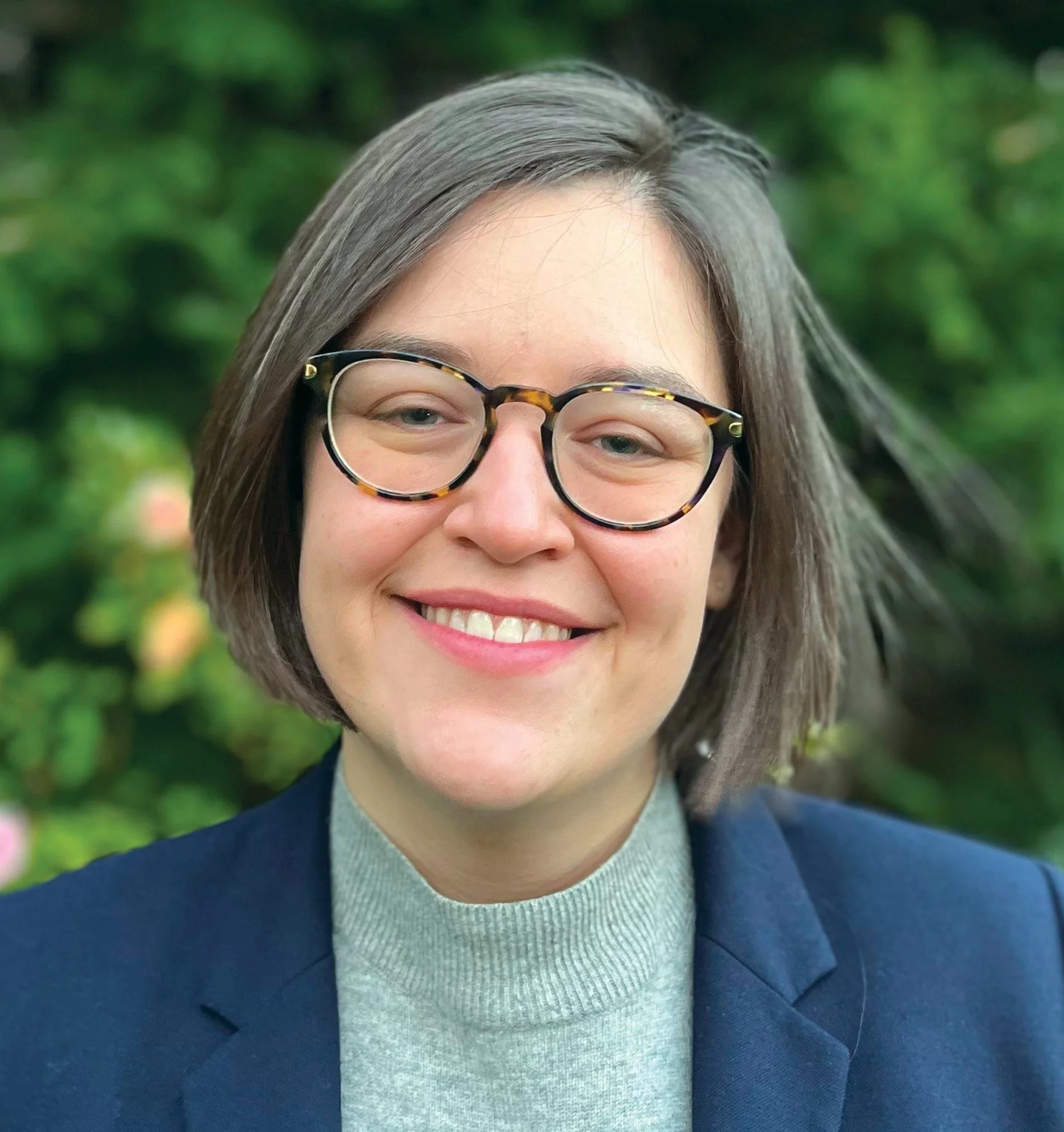 Close-up of a smiling woman with gray hair and tortoise-shell glasses, wearing a navy blazer and gray sweater, outdoors with greenery in the background.