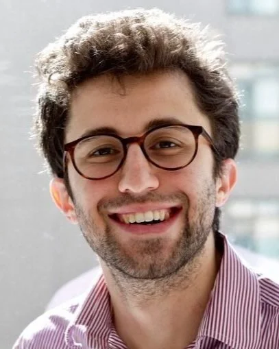 A man with curly hair and glasses smiling, wearing a purple striped shirt.