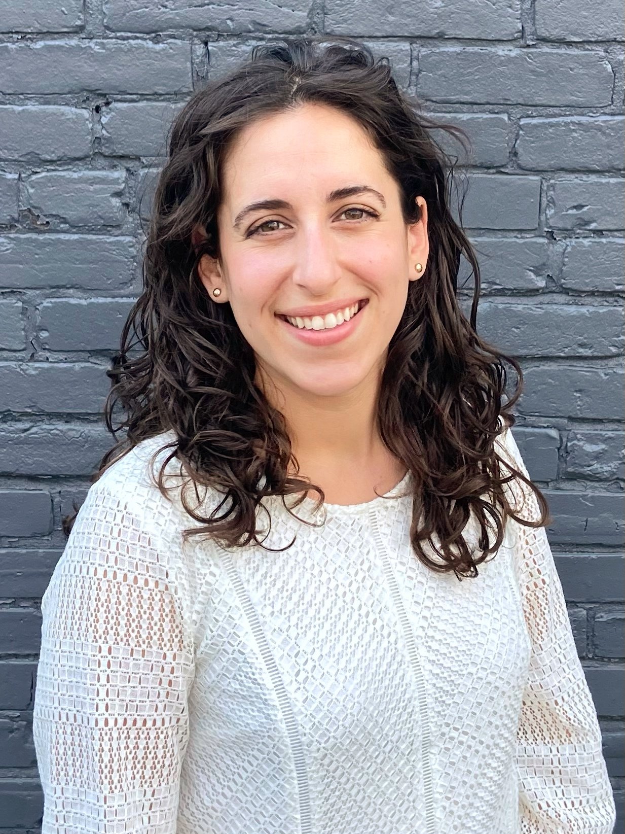 A woman with long, curly dark hair smiling, wearing a white textured top, standing in front of a gray brick wall.