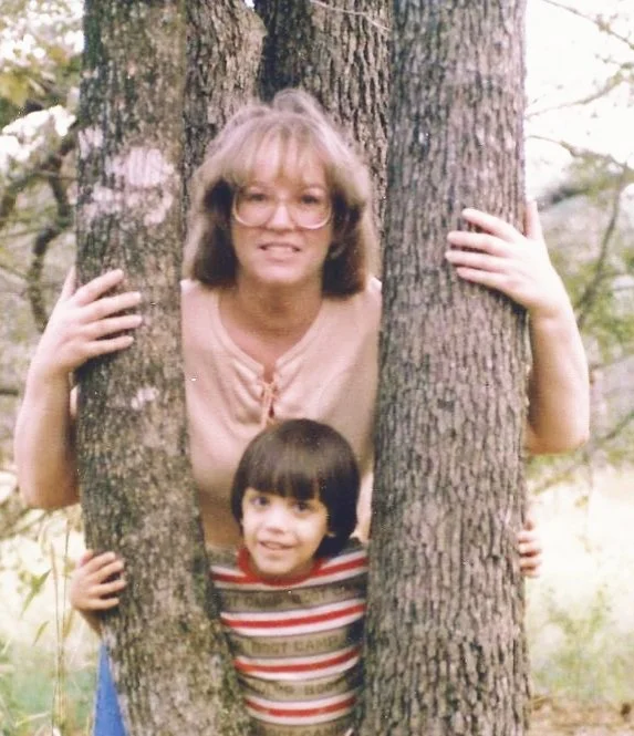 An older woman with glasses and shoulder-length hair standing between two trees, holding onto them with both hands. A young boy with dark hair and wearing a red striped shirt is standing in front of her, smiling. The scene is outdoors with trees and greenery in the background.