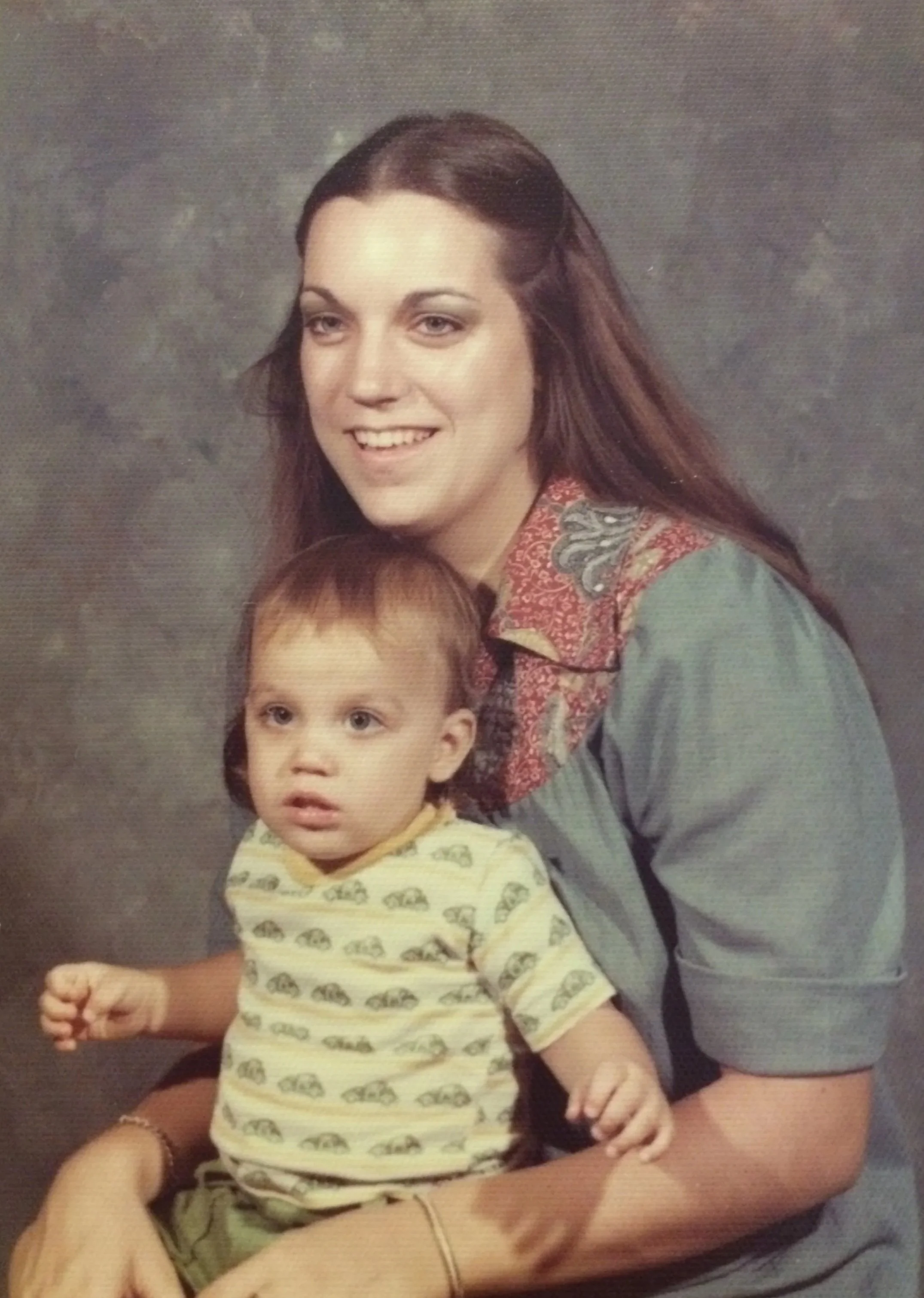 A woman with long brown hair and a gray shirt with floral embroidery, smiling and holding a young boy with short hair and a yellow shirt with animal print, in front of a gray textured background.