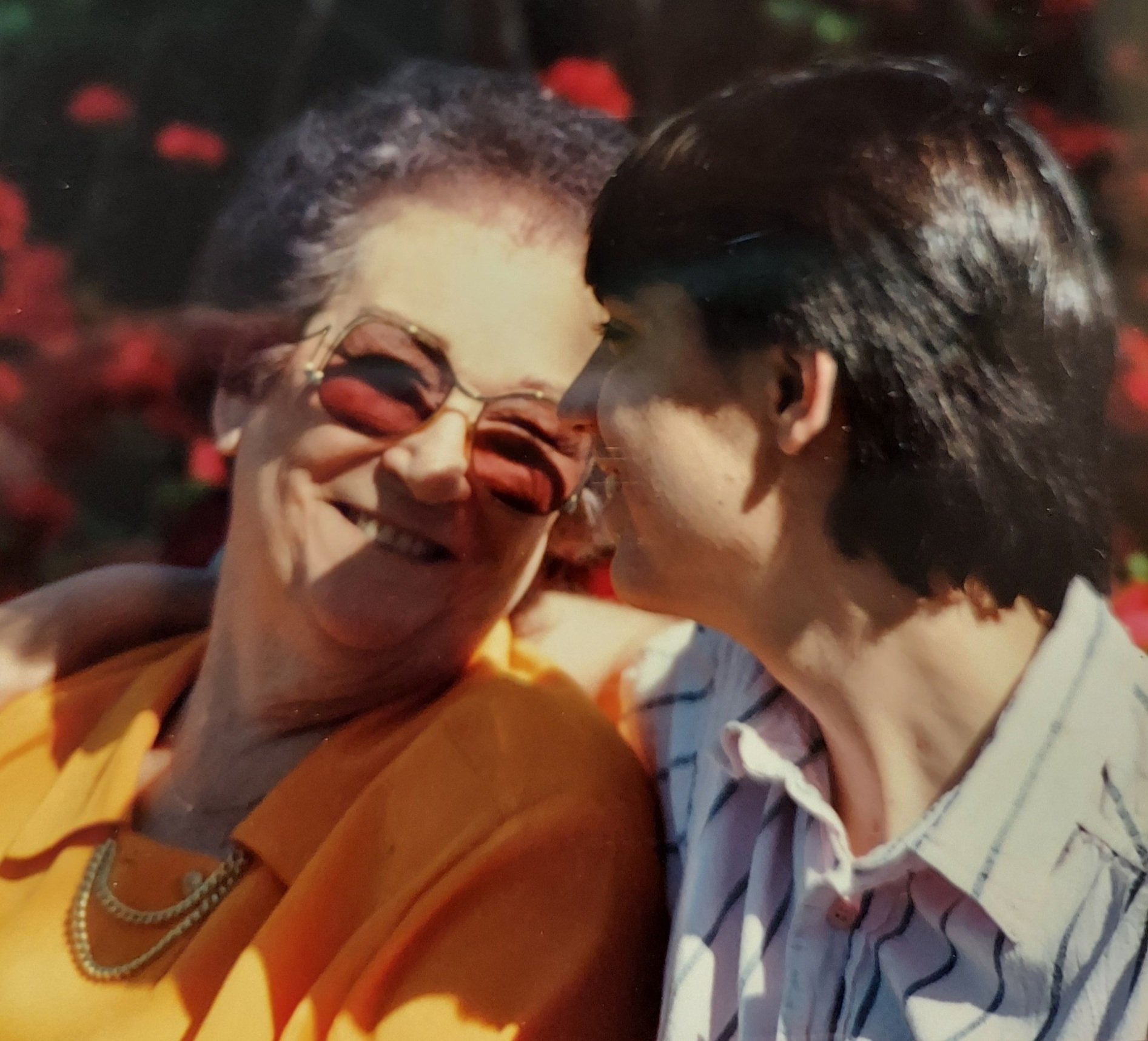 A older woman and middle-aged man sharing a close, happy moment, smiling and looking at each other, with one woman wearing glasses and the other with dark hair, in a warm, outdoor environment.