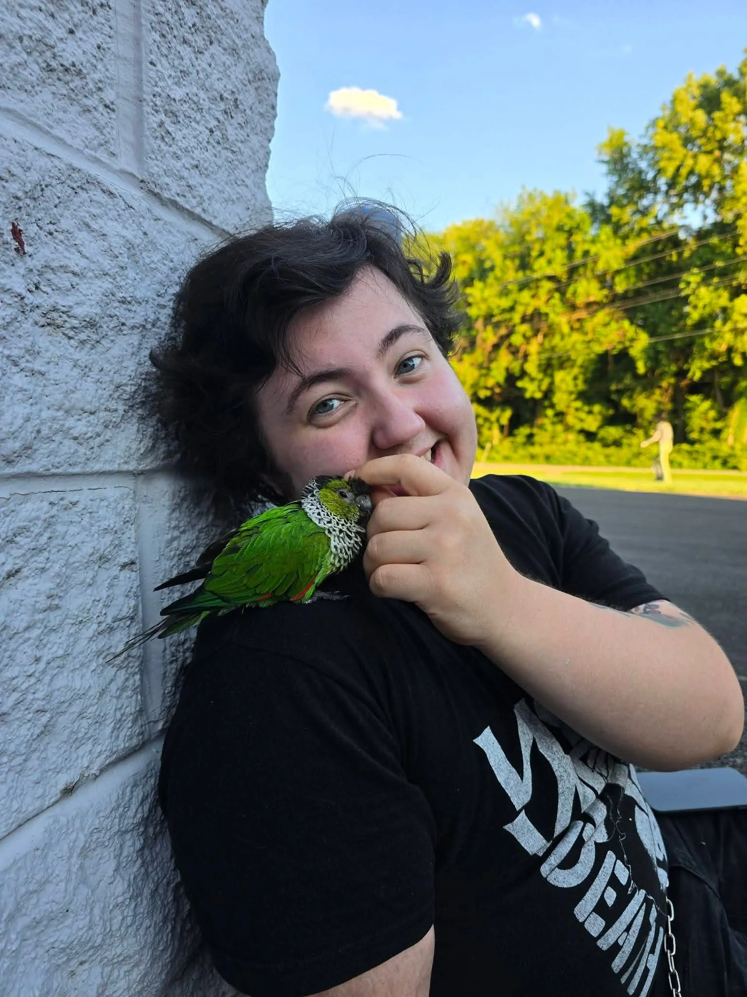 A young person with dark, curly hair and light skin smiling and holding a green parrot with black and white markings on their hand against a backdrop of blue sky and green trees.