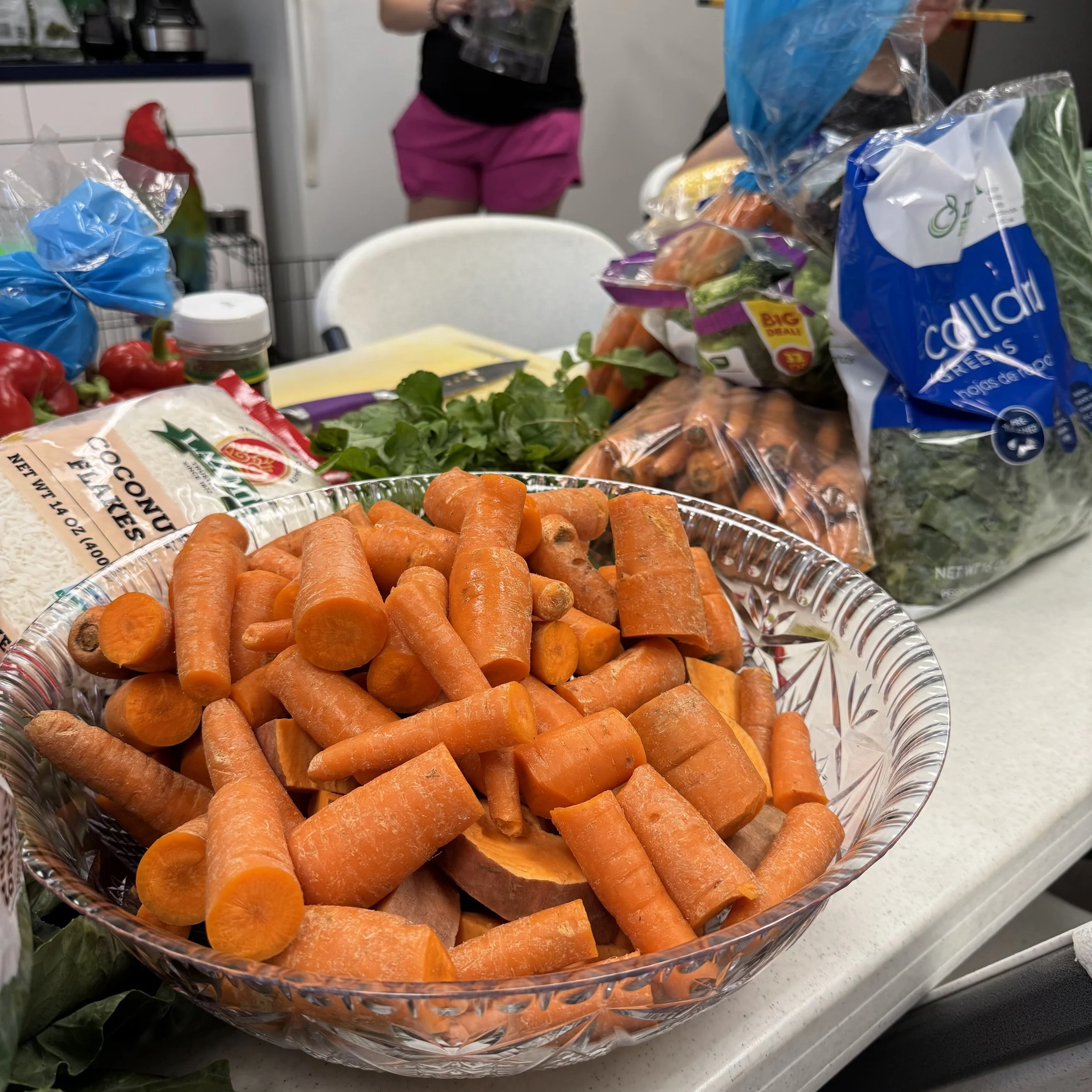 A large glass bowl filled with peeled and cut orange carrots is on a kitchen or dining table. Part of a garden or plant can be seen to the left. In the background, a person wearing black shirt and pink shorts is partially visible, standing next to a 