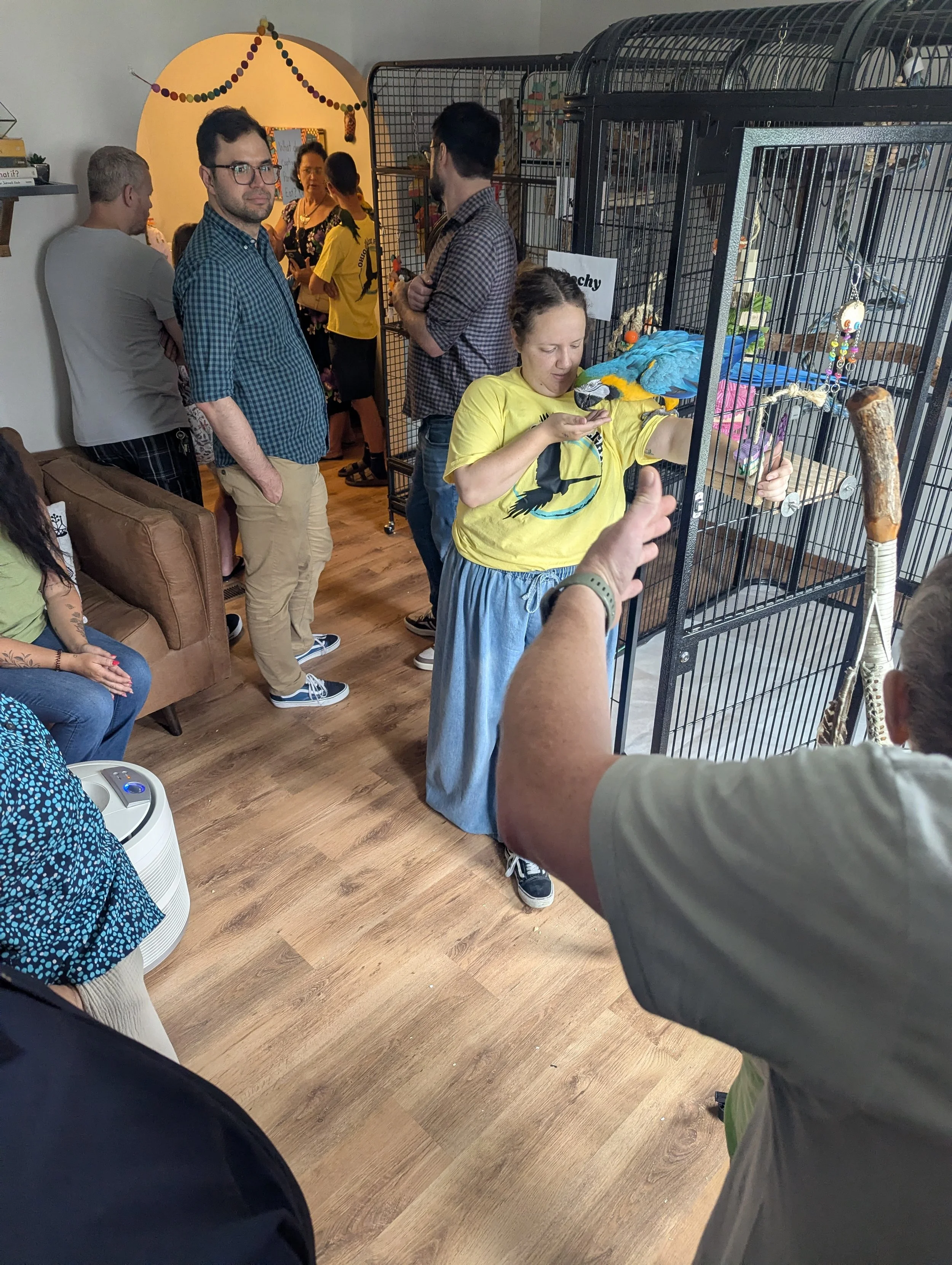 A woman with a parrot on her hand inside a birdcage, with several people in line behind, some looking at the bird and others talking, at an indoor pet event.