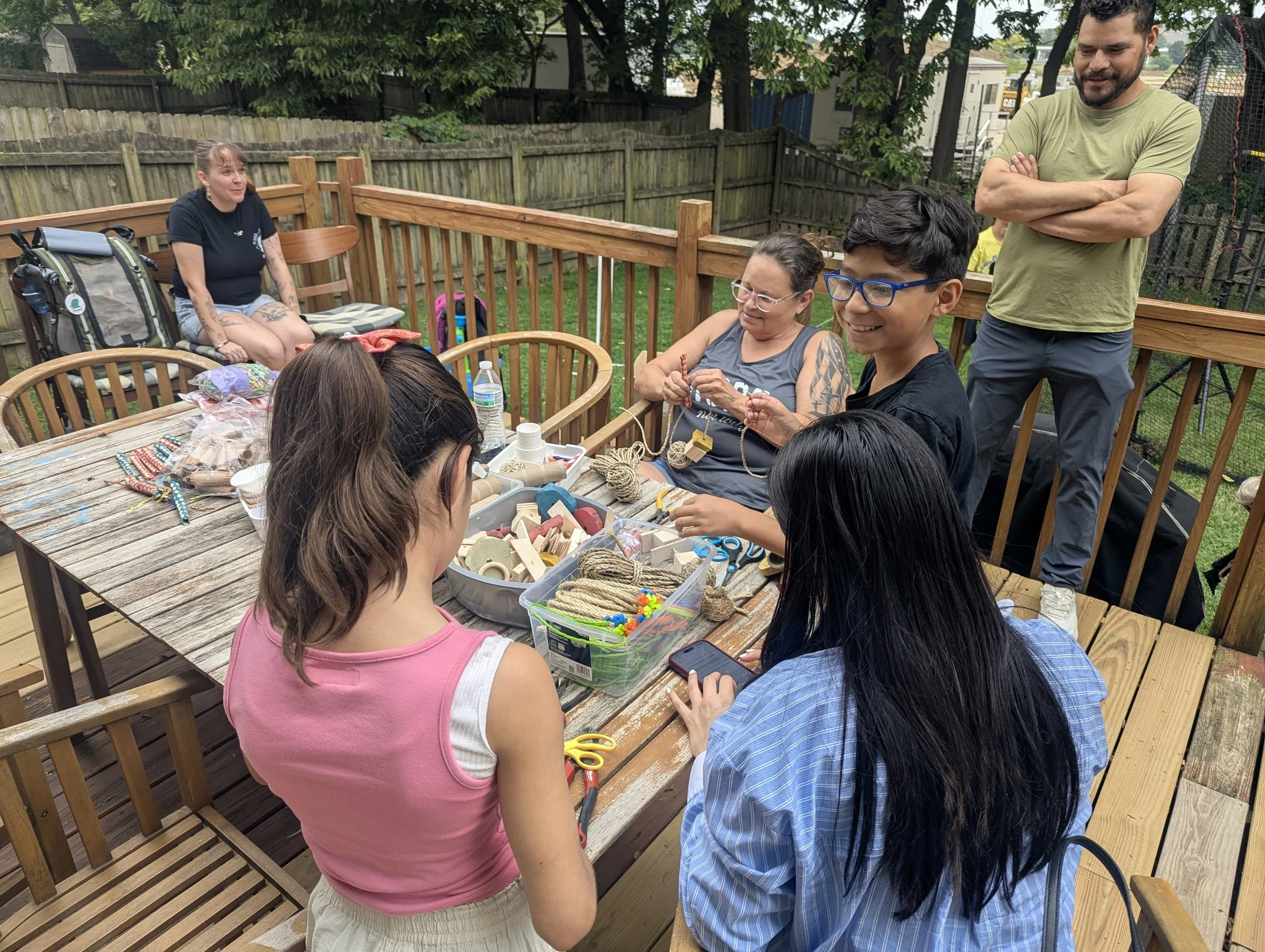 A group of people sitting and standing around a wooden table outdoors, engaged in craft activities, with supplies like rope, scissors, and decorations on the table, in a backyard with a wooden fence and green trees.