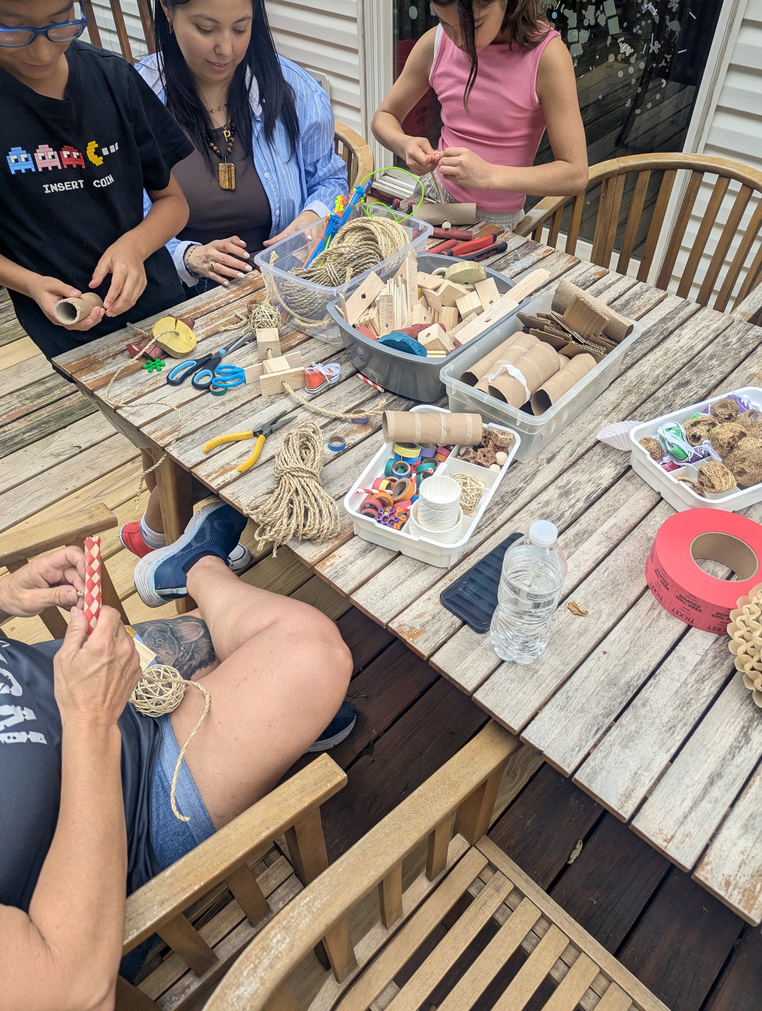 People crafting with various materials, including wood, string, and cardboard, at an outdoor wooden table. There are scissors, tape, and other supplies on the table.