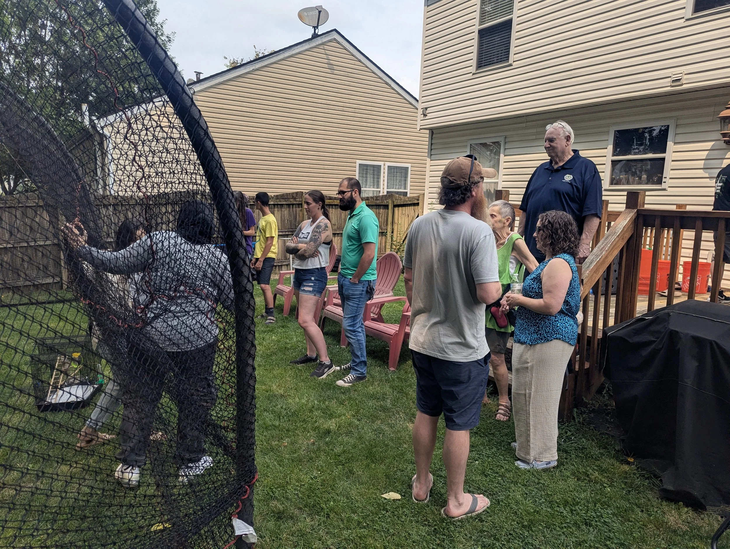 A backyard gathering with multiple people standing and chatting, some seated on pink chairs, with a house and a wooden deck in the background, and a forested area beyond the fence.