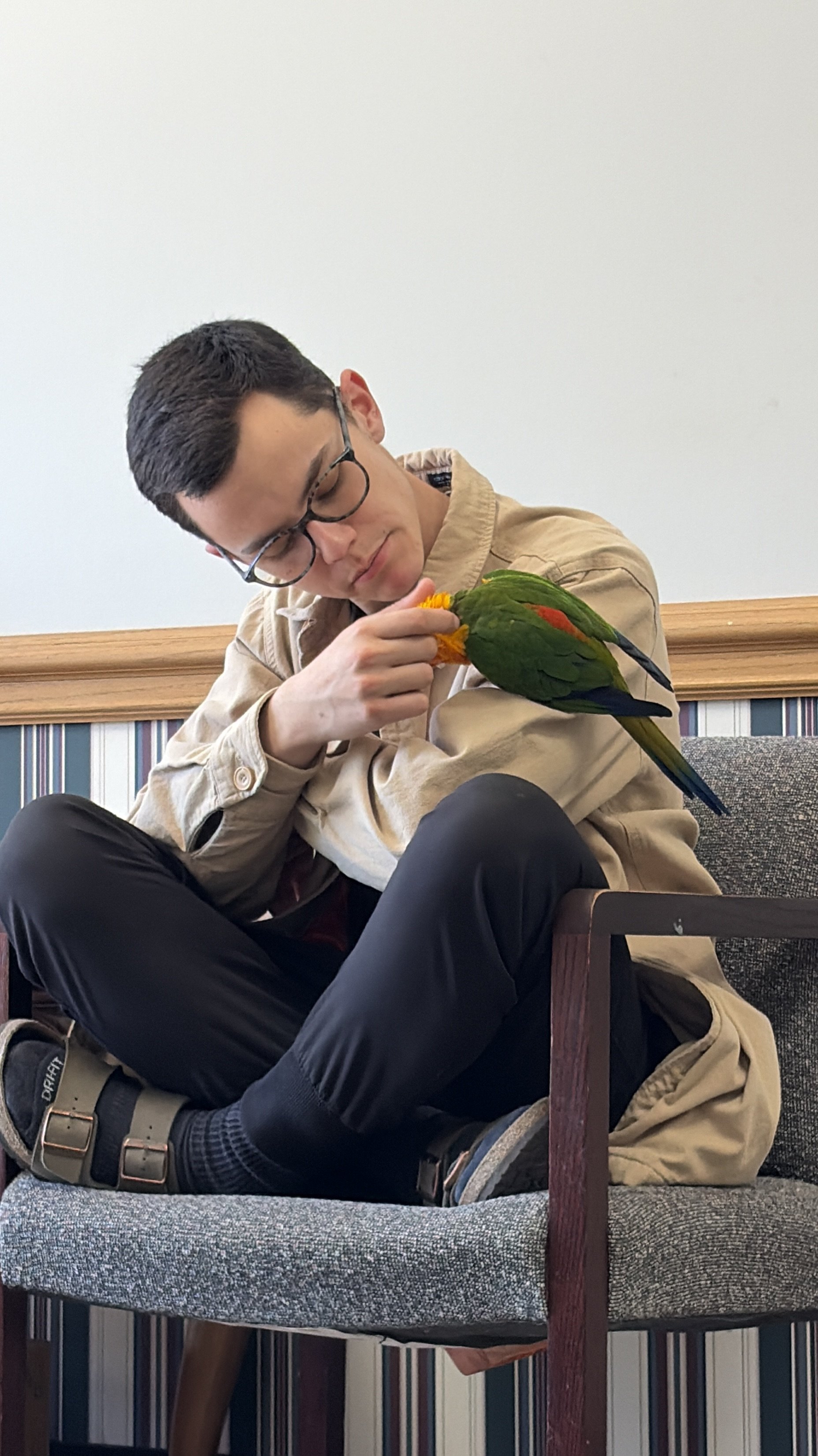 A young man with dark hair, glasses, and a beige jacket sitting on a gray upholstered bench, holding a colorful green and red bird on his hand, indoors with a plain wall in the background.