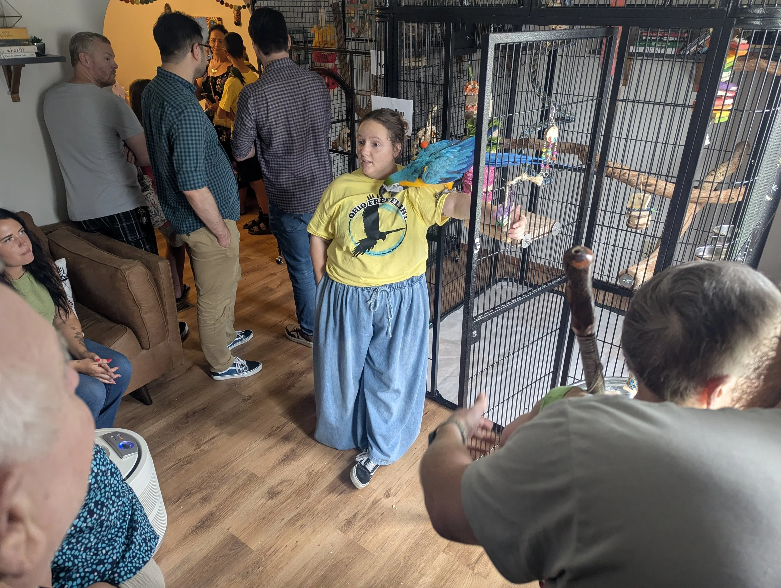 A group of people gathered around a large cage with colorful birds inside at an indoor event.