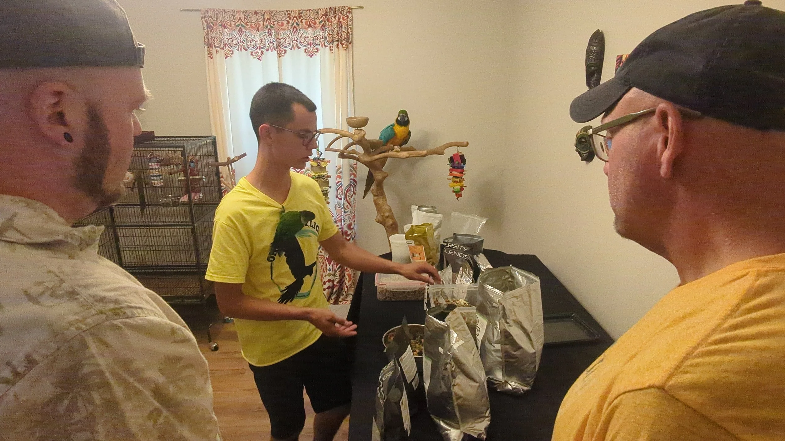 Four people are gathered around a table with snacks and food in a room. One person is in the background near a bird on a perch and a cage. The room has a window with colorful curtains.
