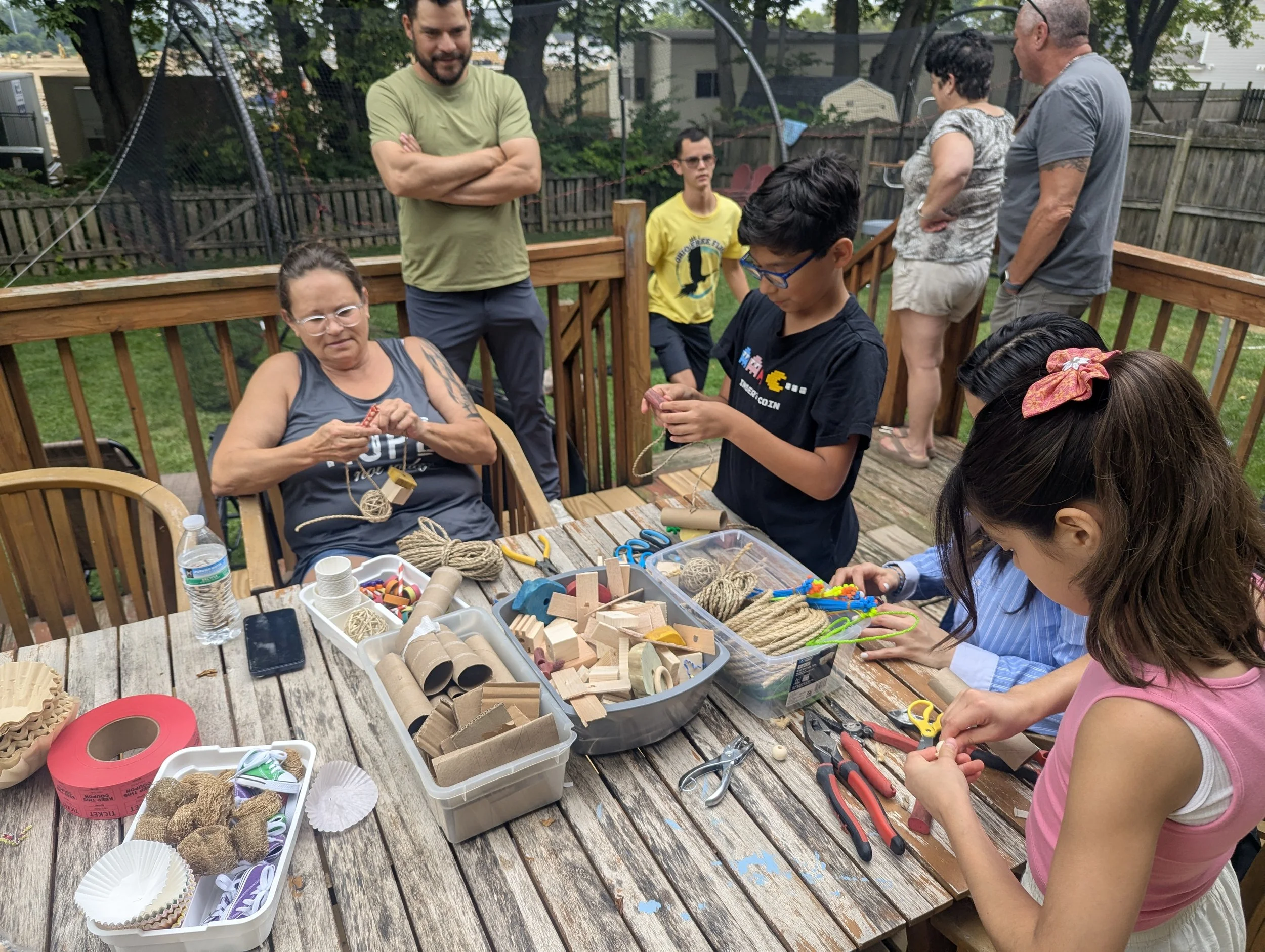 A group of people are gathered on a wooden deck, engaging in arts and crafts activities with various crafting supplies such as yarn, scissors, and wooden blocks. Some are working on projects, while others are standing and observing. There are trees a