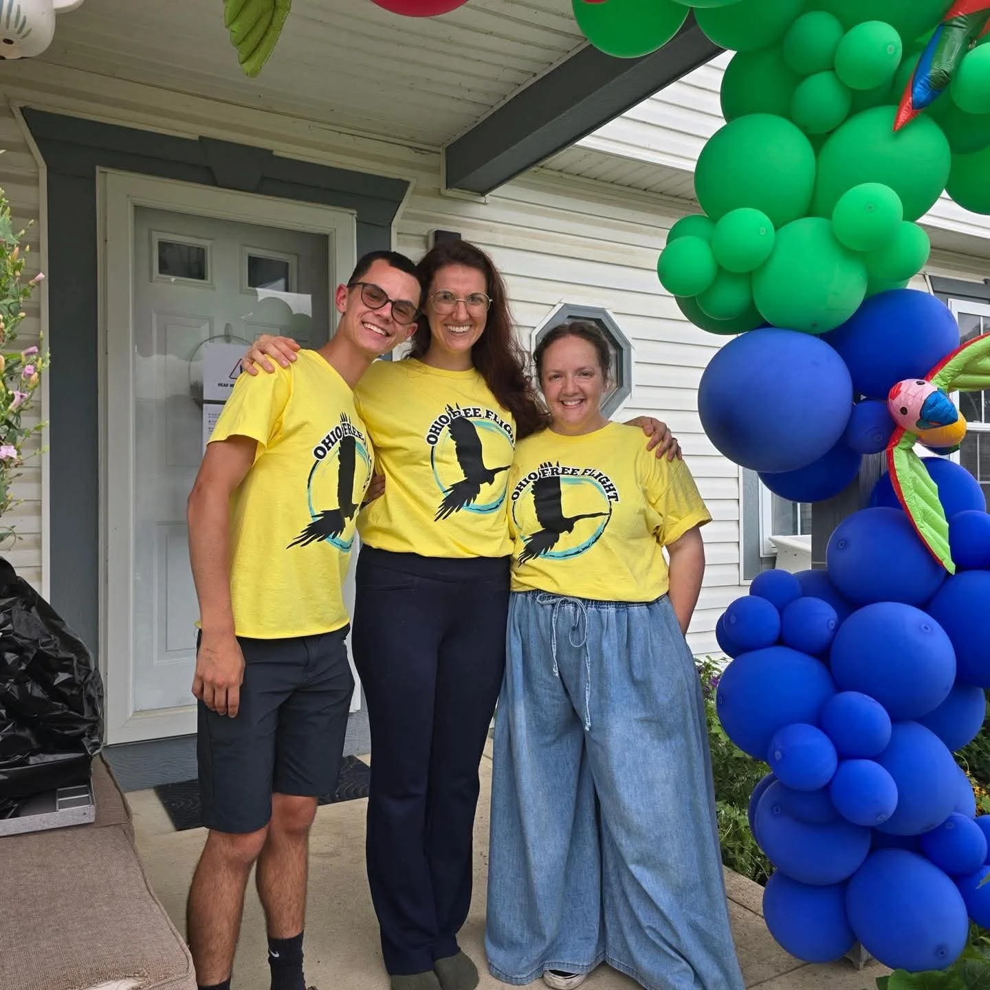 Three people standing on a porch next to a balloon arch, all wearing yellow shirts with a black bird silhouette and the text 'Ohio Free Flight'.
