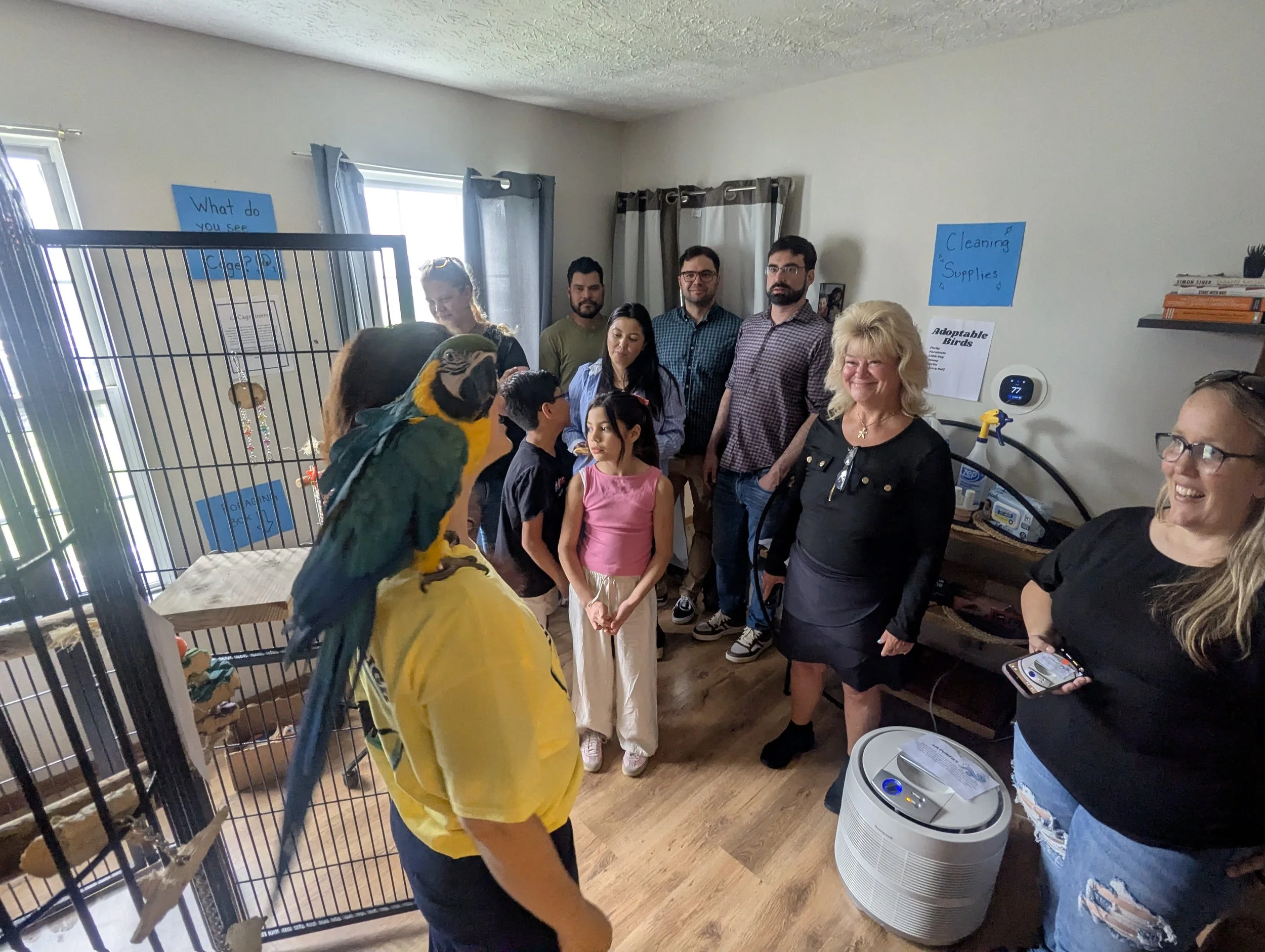 A group of people gathered inside a room with a bird in the foreground. The room has windows with light curtains, bird cages, and signs on the wall including one that reads 'Cleaning Supplies.' Several adults and children are observing the bird and s