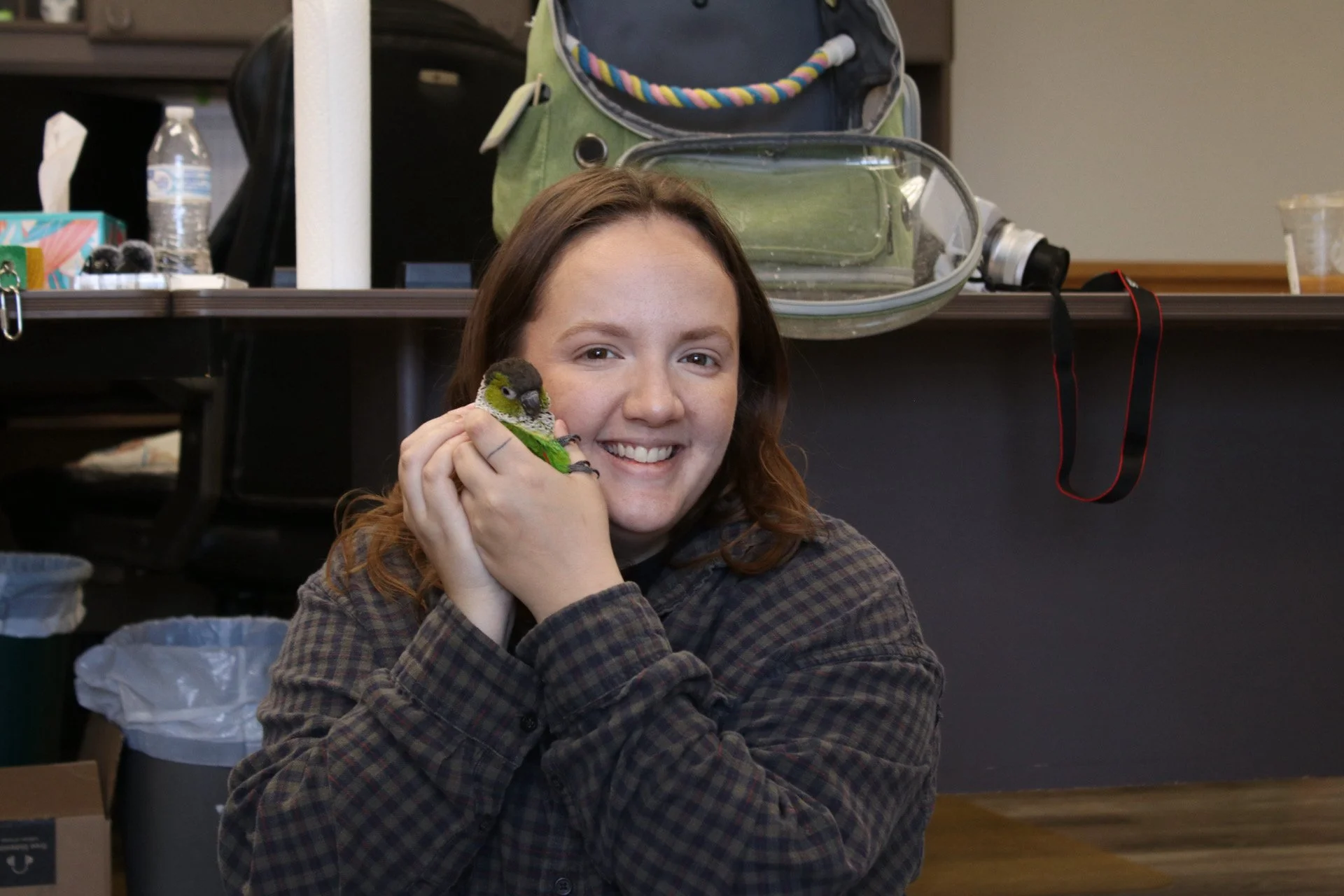 A woman with brown hair smiling while holding a small green and black parrot. He is a Black Capped Conure