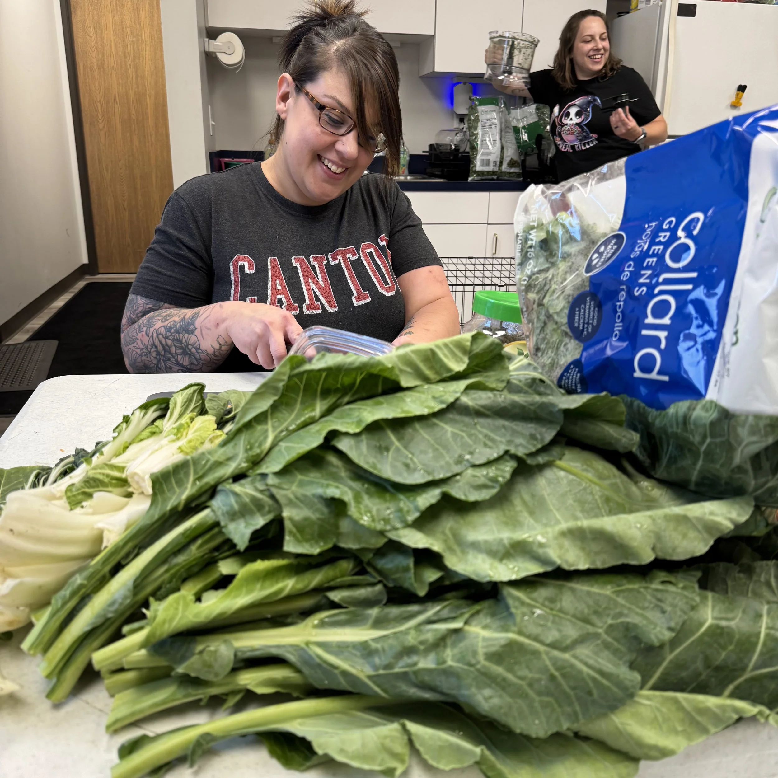 Two women in a kitchen with one slicing lettuce and the other holding a glass jar, surrounded by salad ingredients and a large packet of romaine lettuce.