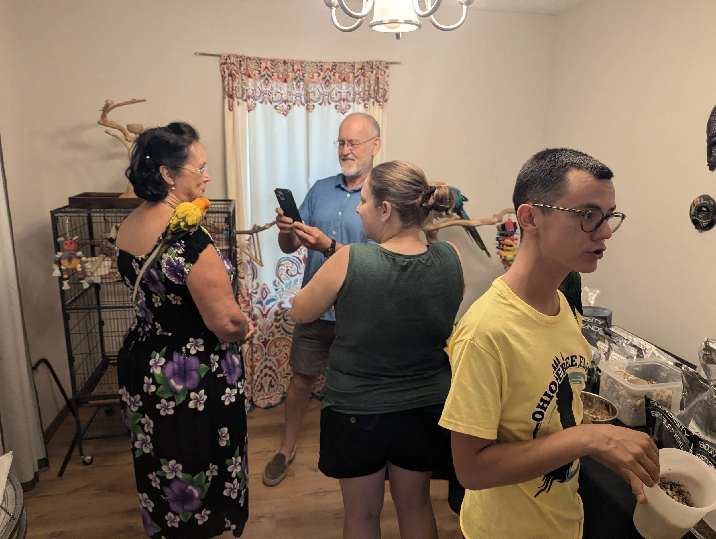 Group of people in a room, a woman with a bird on her shoulder, a man in a blue shirt showing something on his phone, a woman holding a drink, and a young man with glasses at a table with snacks.