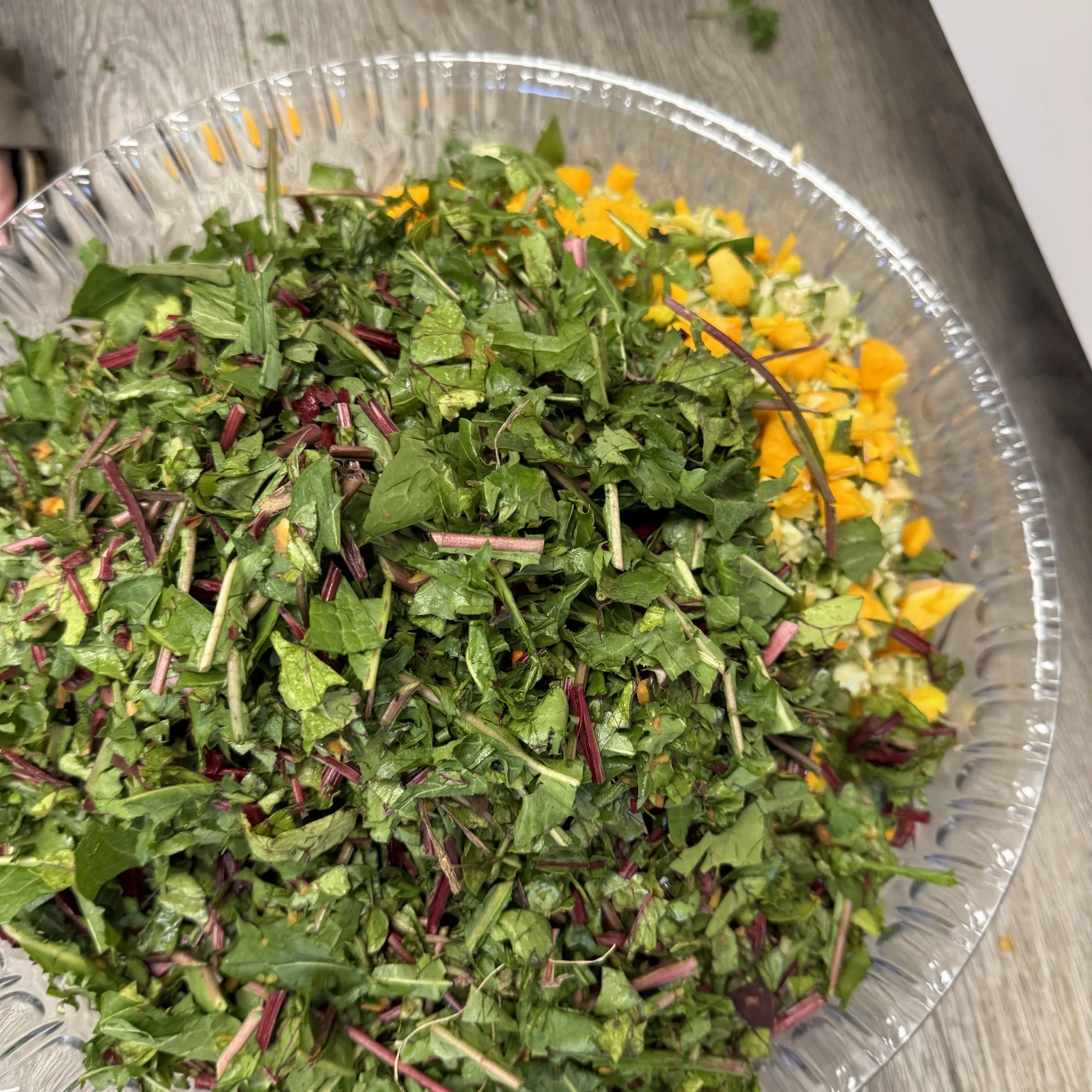 Close-up of a glass bowl filled with mixed chopped leafy greens and shredded orange and yellow vegetables, placed on a wooden surface.