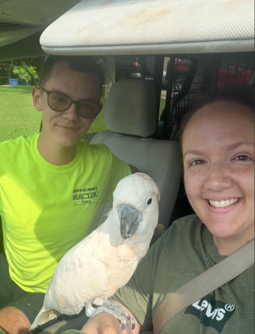 A woman and a young man sitting inside a vehicle with a white cockatoo perched on the woman's arm, both smiling at the camera.