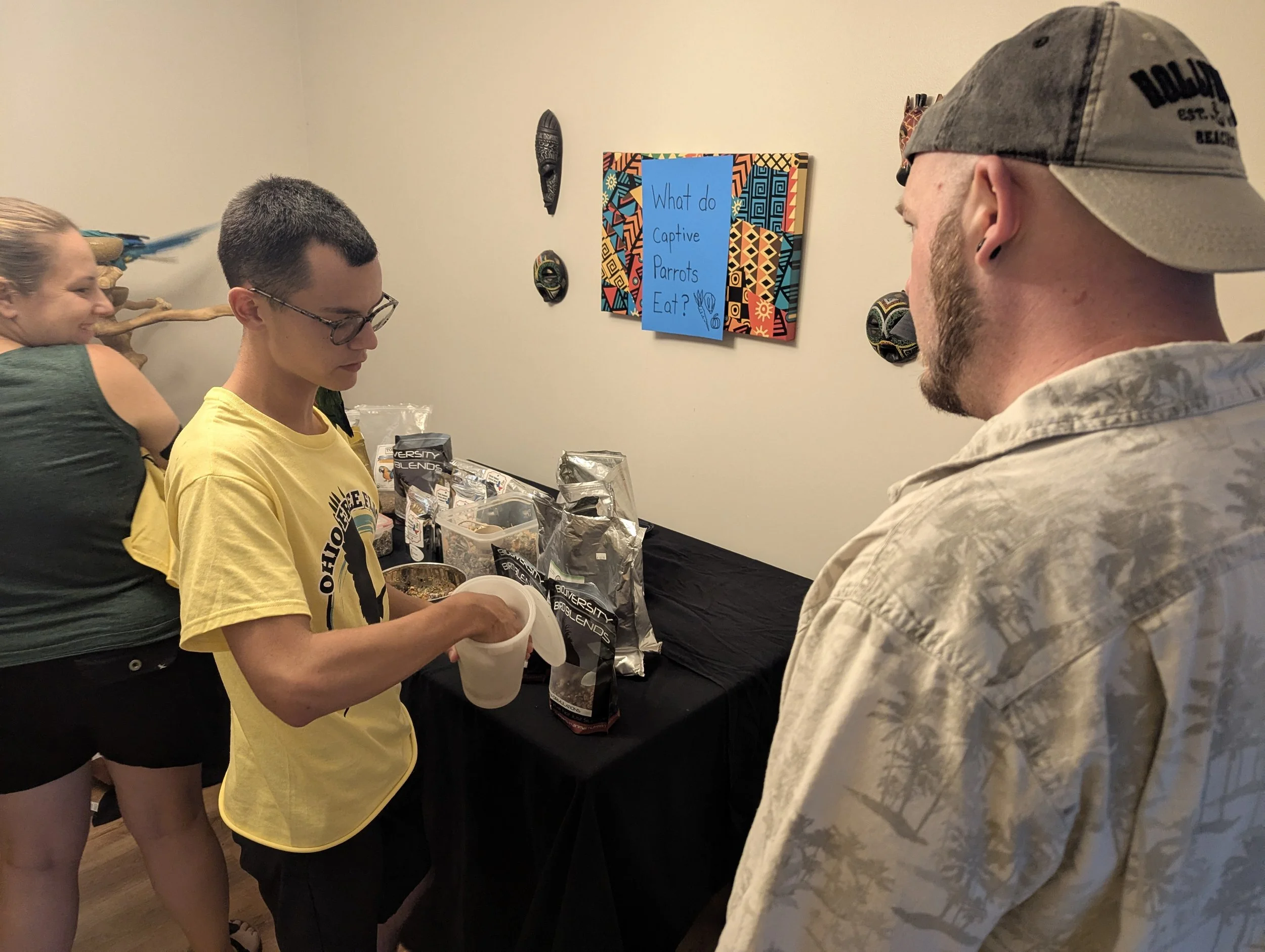 Three people standing indoors around a table with snacks. A woman with her hair tied up and a woman with glasses, both in T-shirts, are looking at a man in a camouflage shirt and cap. A colorful sign on the wall reads, "What do Captive Parrots Eat?"
