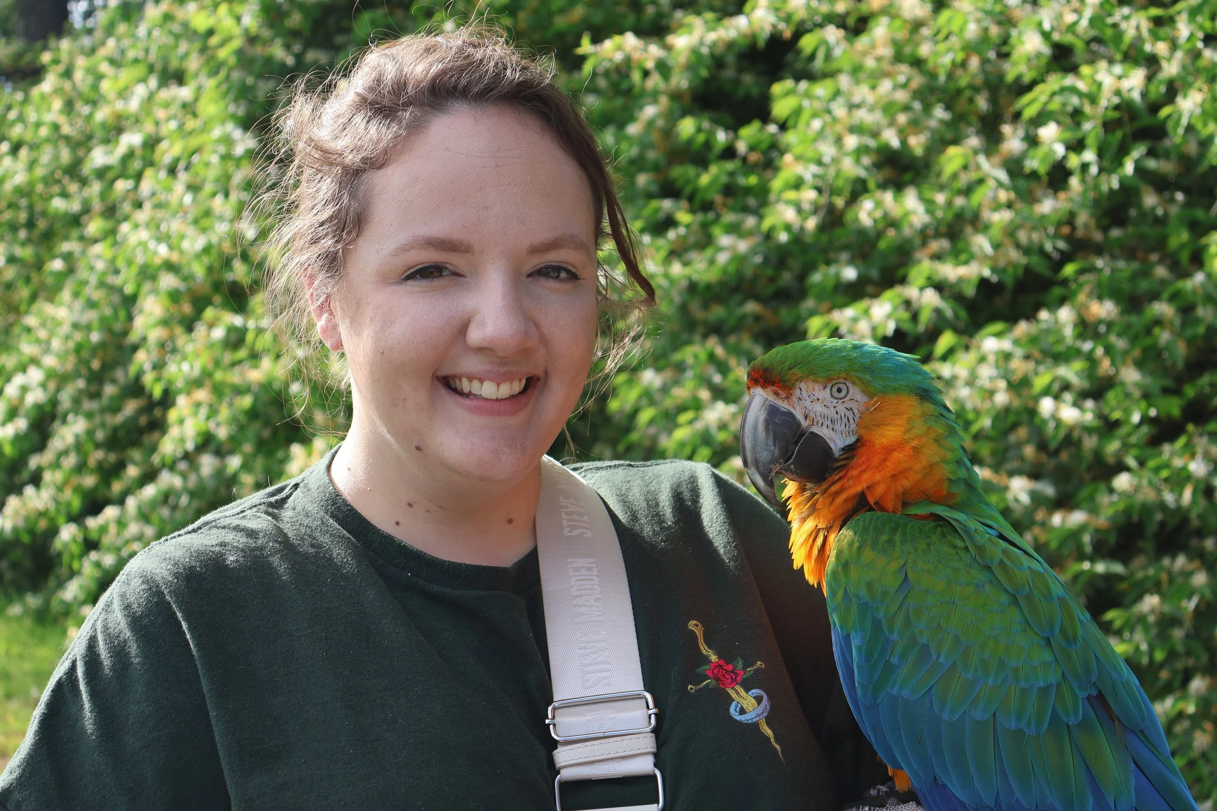 A woman smiling with a colorful macaw perched on her shoulder, outdoors with greenery in the background.
