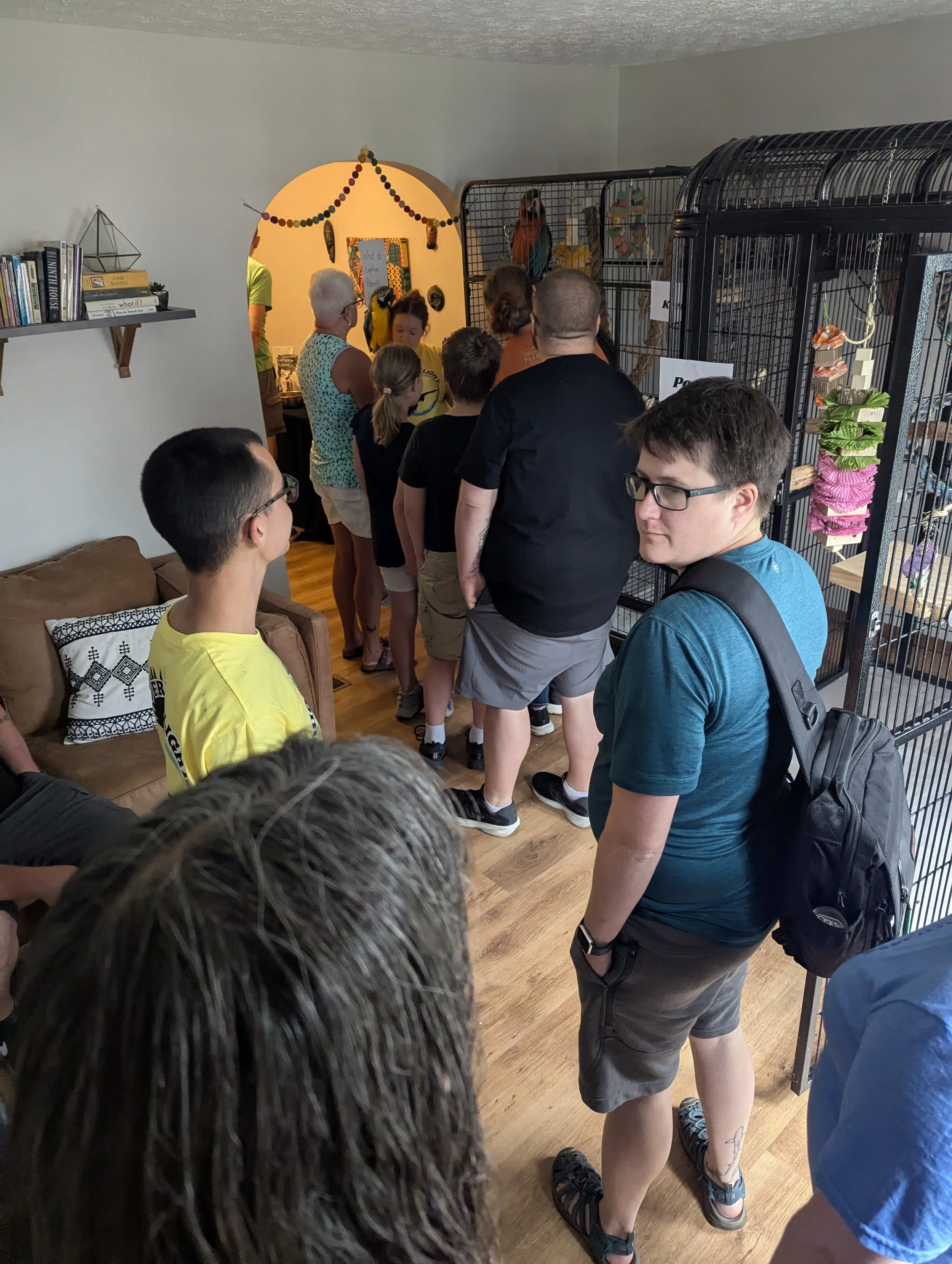 A group of people standing in line indoors, waiting behind metal cages with various toys and items inside. The room has wooden flooring and decorative wall hangings.