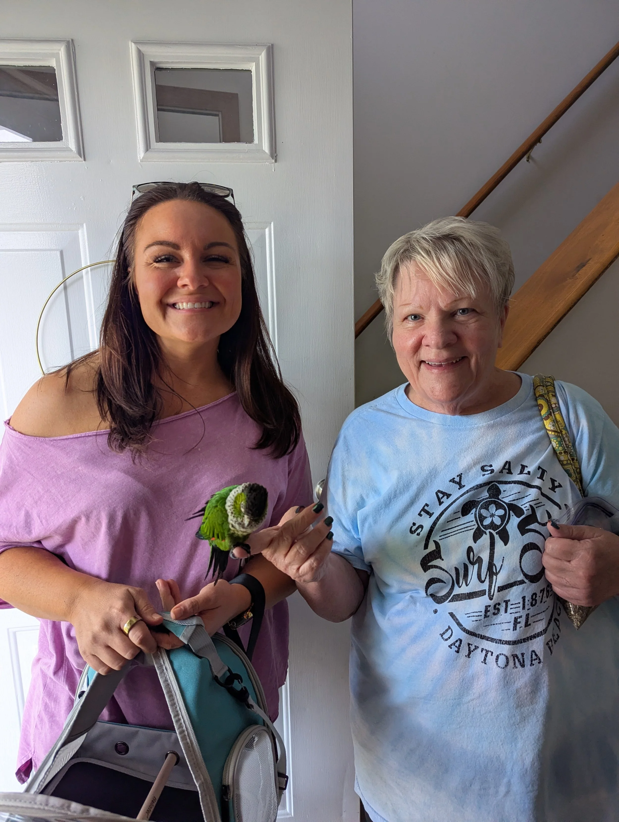 Two women standing indoors near a staircase and a door, smiling and holding small parrots, one with green and black feathers and the other with blue and black feathers.