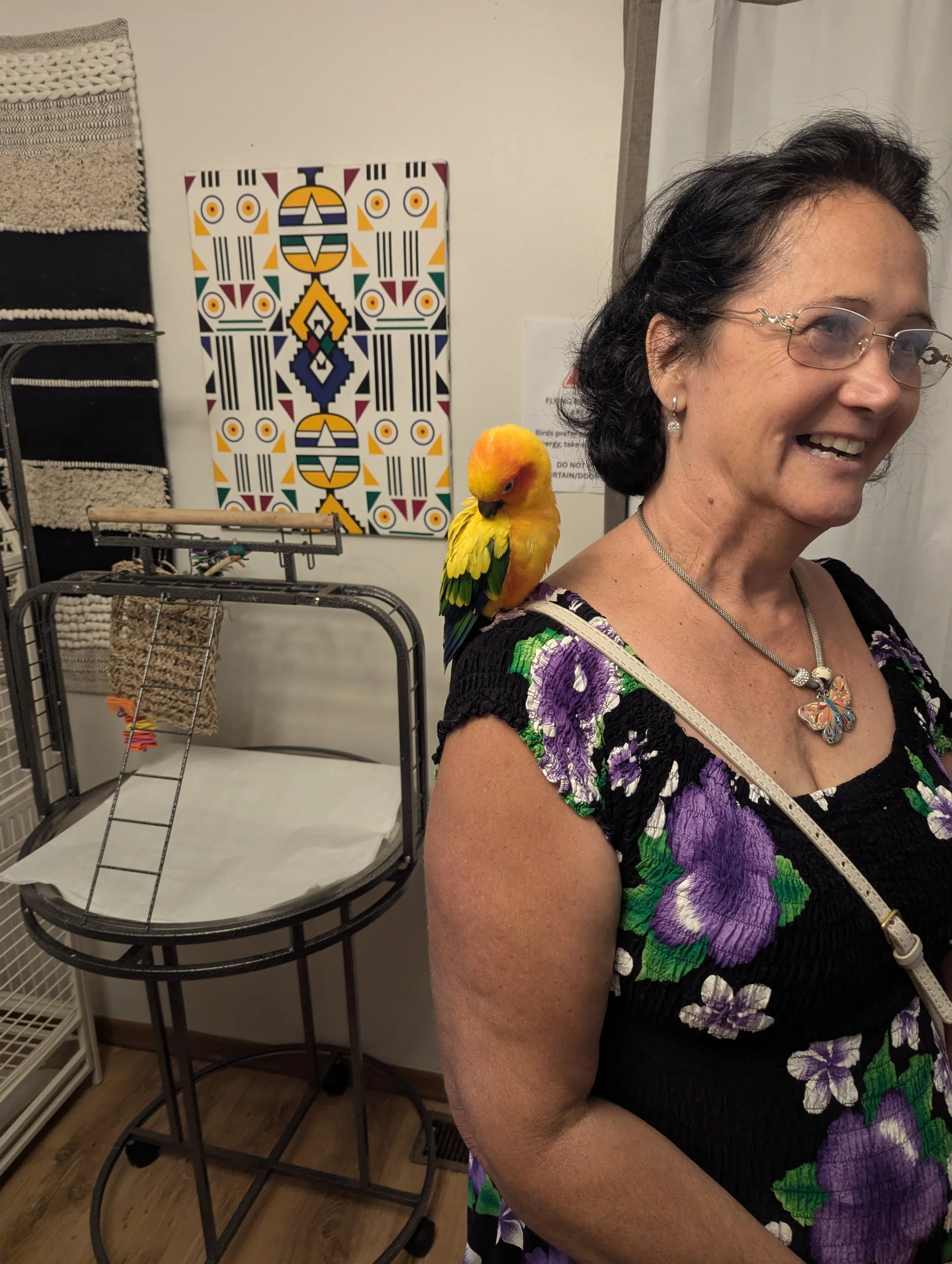 A woman with glasses and earrings smiling, standing next to a colorful geometric art piece on a white wall, with a bird perched on her shoulder, partially visible in the foreground.