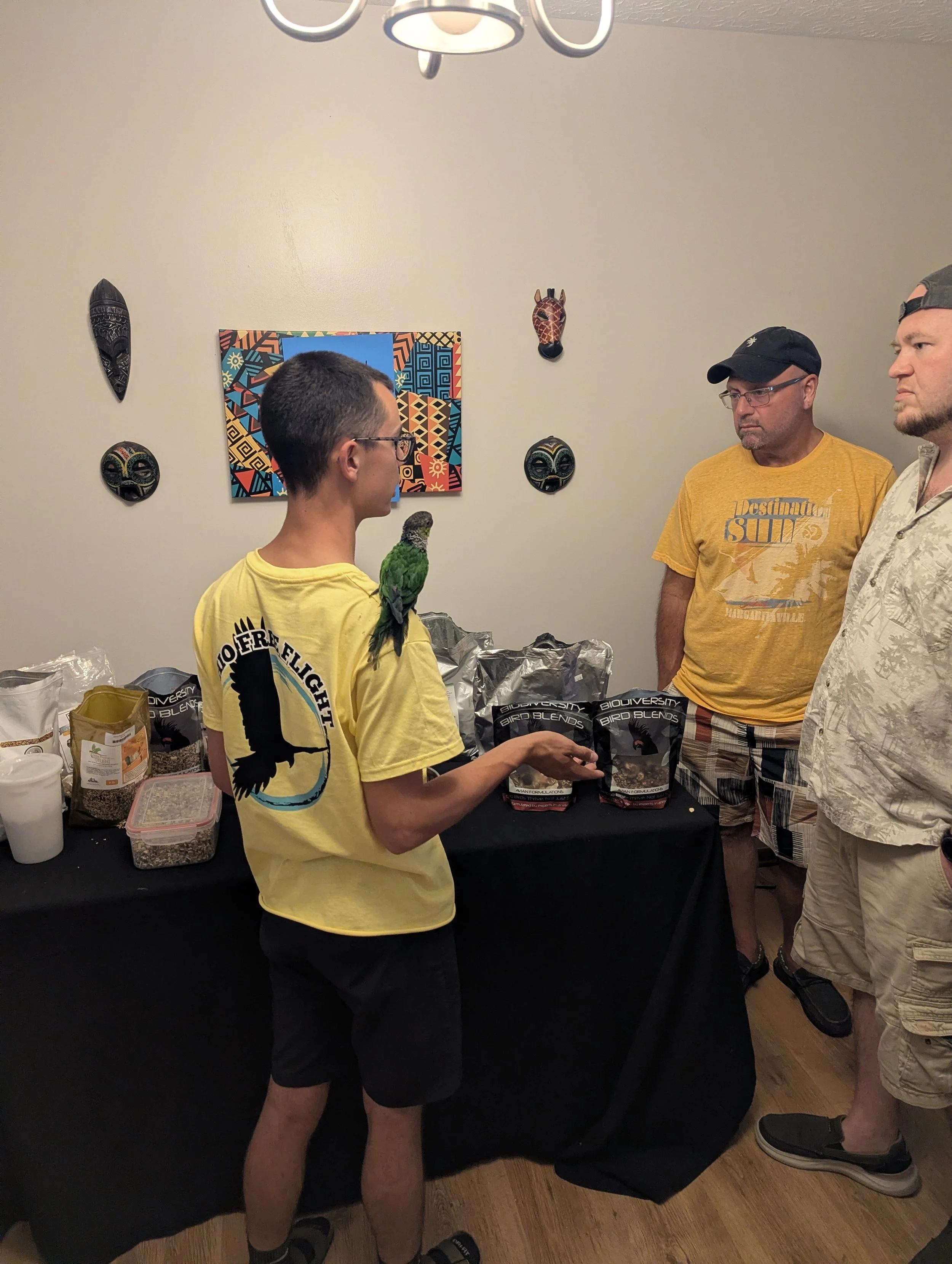 Three men are standing around a table with various bags of bird seed and other supplies. One man is holding a small green and yellow bird, possibly a parrot, on his arm. The background features a colorful artwork and African masks on the wall.