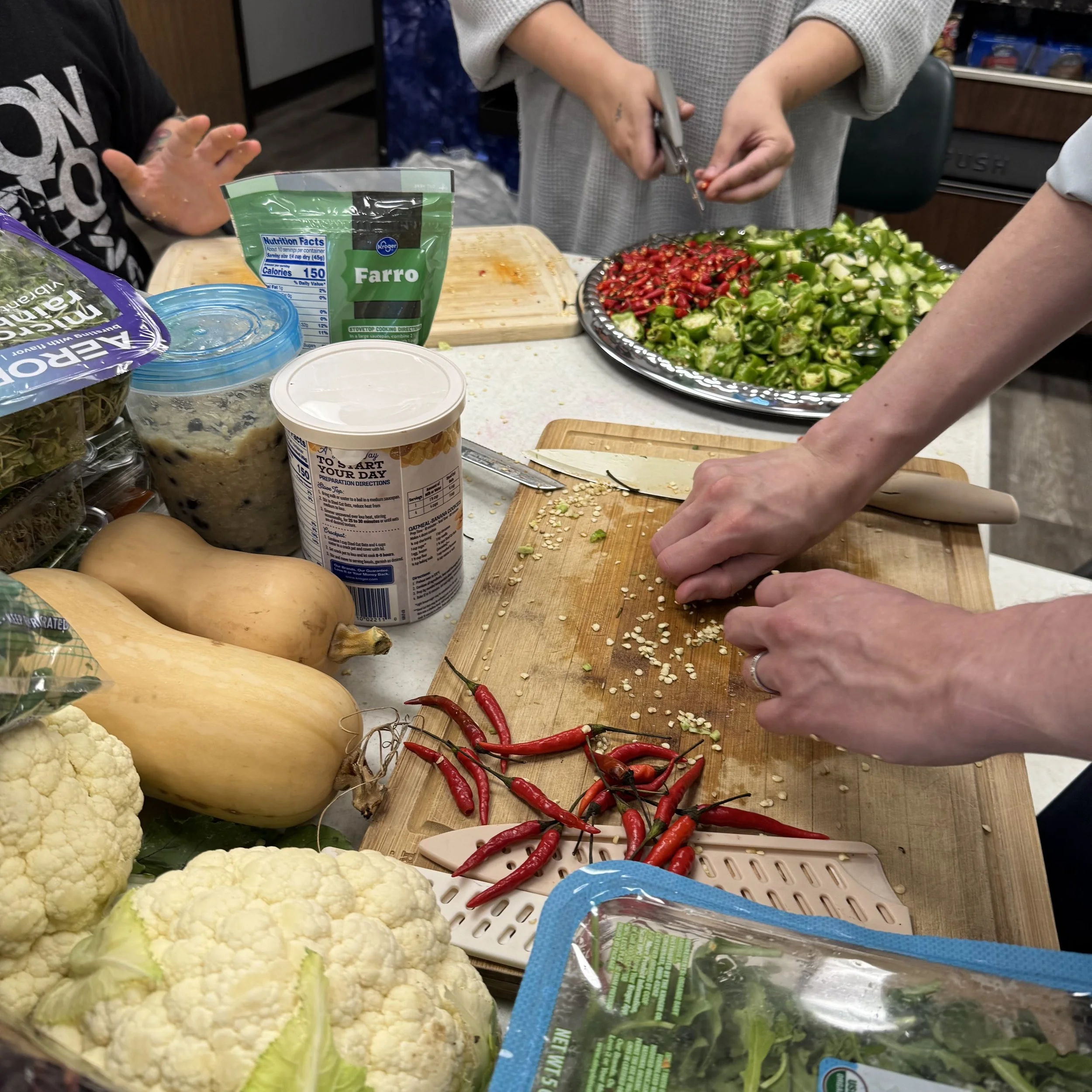 People preparing vegetables in a kitchen, with cauliflower, red chili peppers, and leafy greens on the counter.
