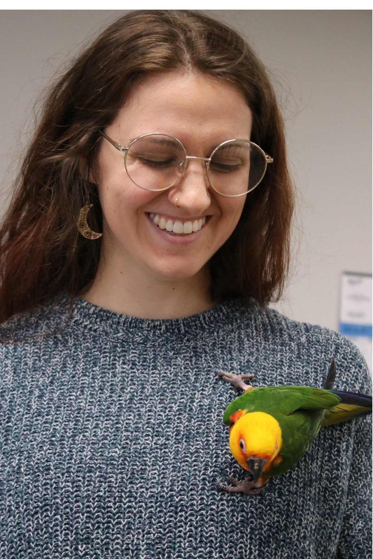 A young woman with brown hair, glasses, and a nose ring, smiling and looking at a colorful parrot perched on her hand. He is a Jenday Conure.