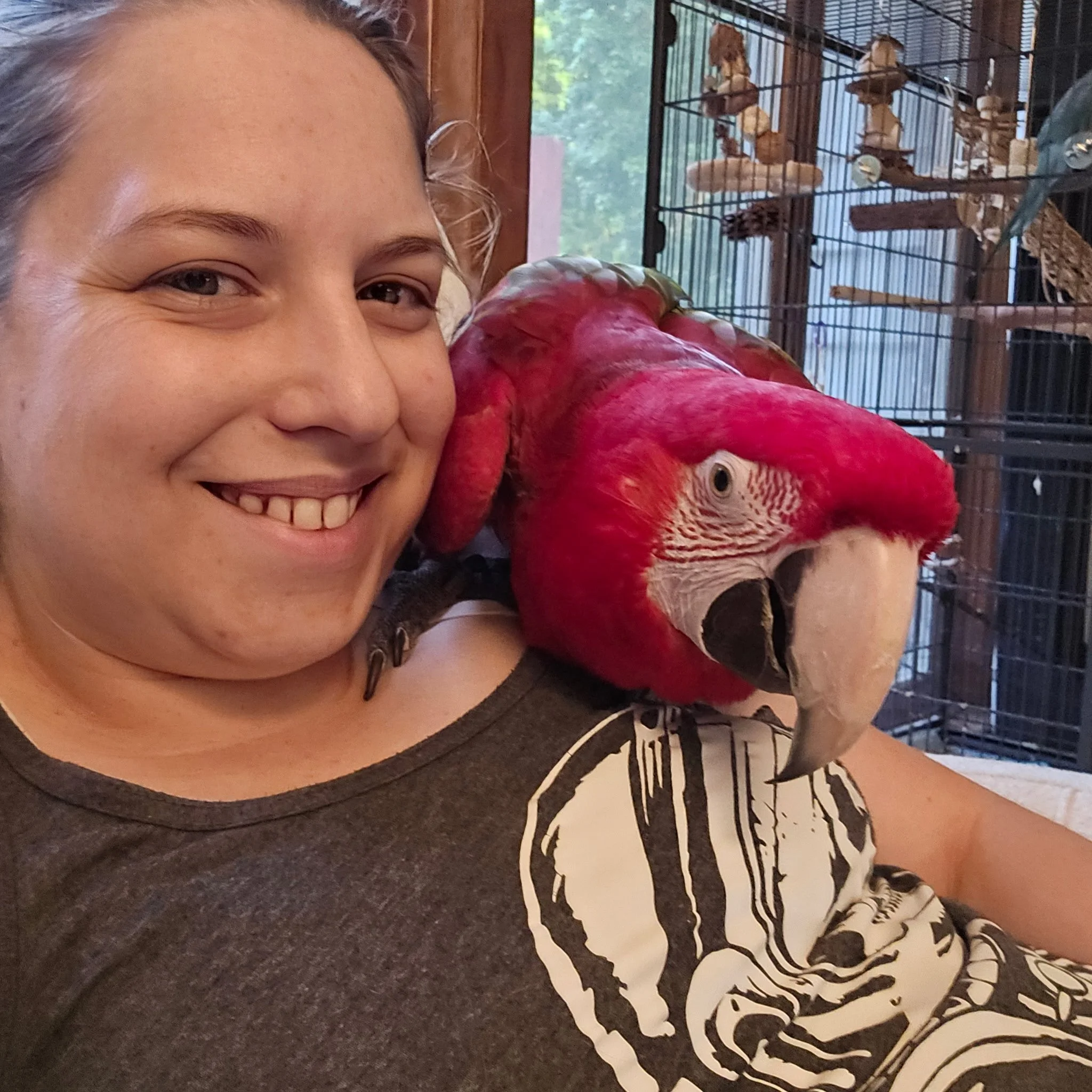 A woman smiling and leaning against a red parrot in front of a cage filled with various birds.
