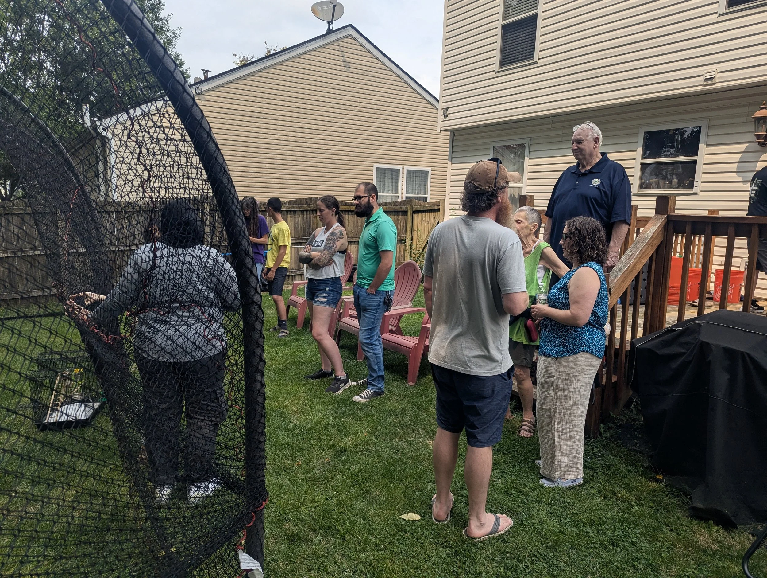 A group of people gathered in a backyard, some standing and talking, with chairs and a wooden deck visible. The yard is enclosed by a wooden fence, and there are houses in the background. The sky is cloudy, and the scene appears to be at a social gat
