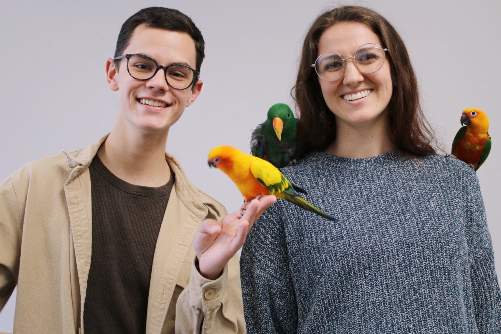 A man and woman smiling at the camera with colorful parrots perched on their arms and shoulders, standing against a plain light gray background. The birds are Sun Conure, Jenday Conure, and Eclectus.