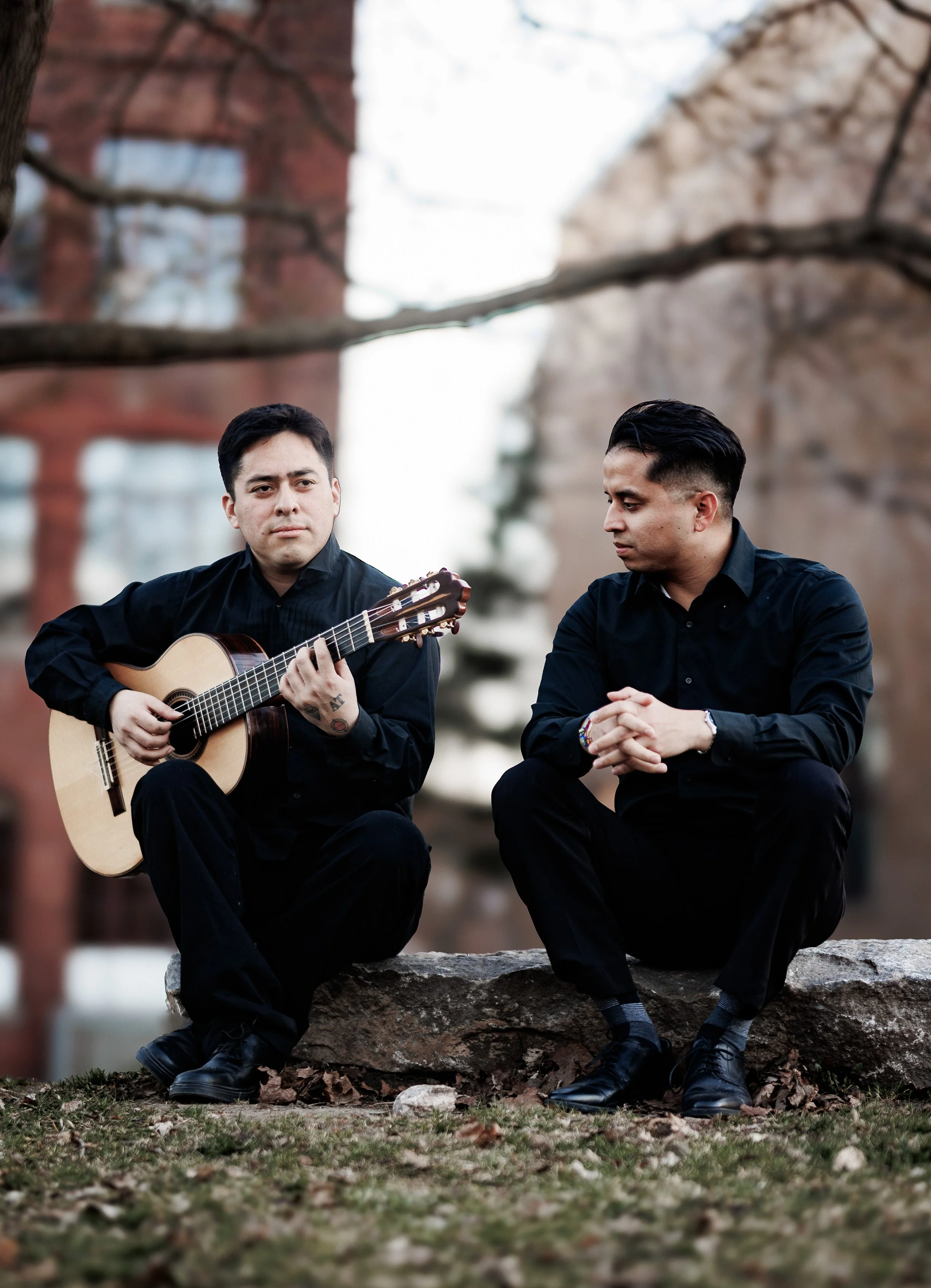 Two young men sitting outdoors on a rock, one playing guitar and the other listening, with trees and buildings in the background.