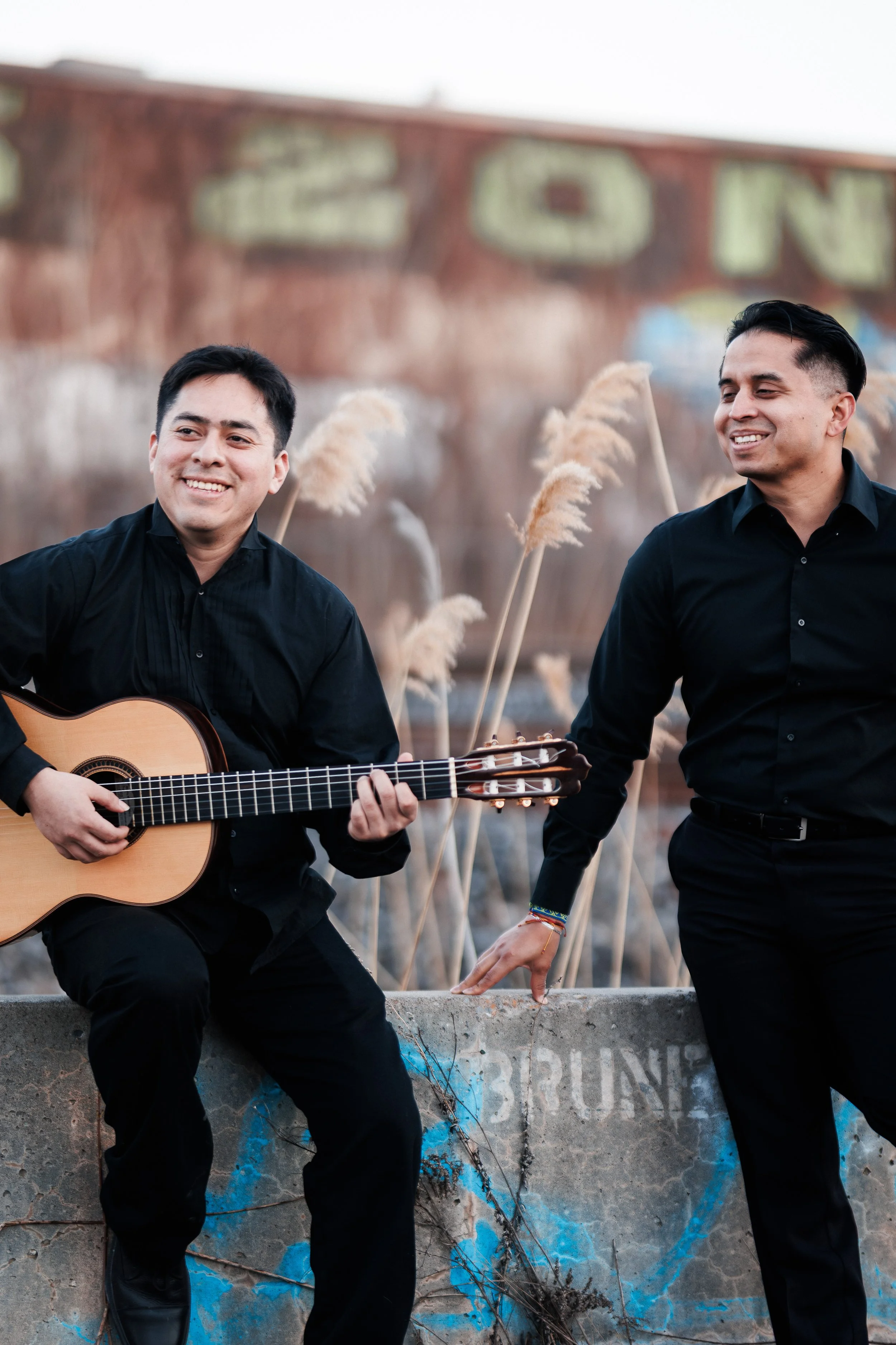 Two men in black shirts outdoors with tall grass and graffiti on a wall in the background. One man is playing guitar and smiling, the other is leaning on a concrete ledge and smiling.