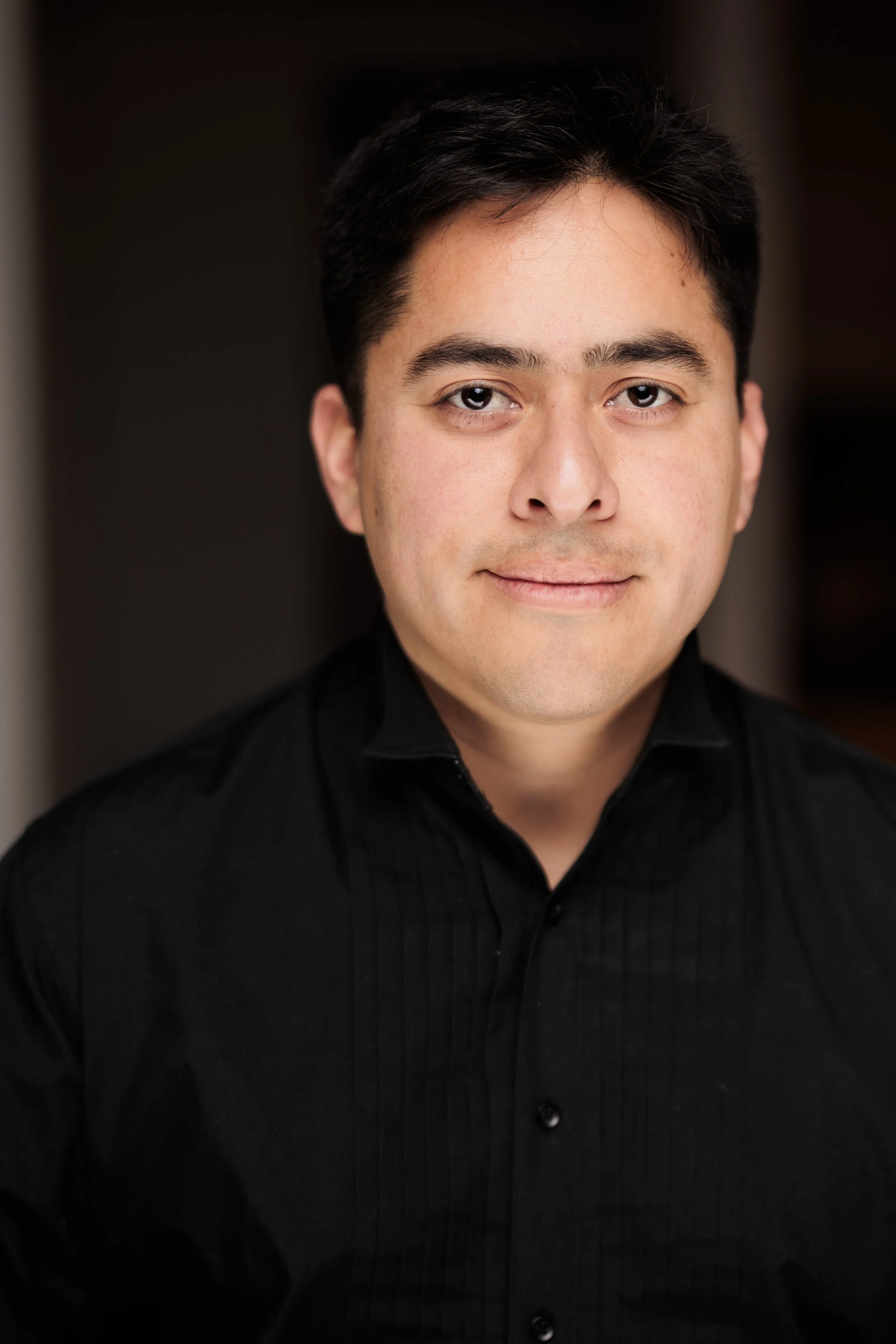 Close-up of a young man with dark hair and light skin, wearing a black shirt, looking at the camera with a neutral expression.