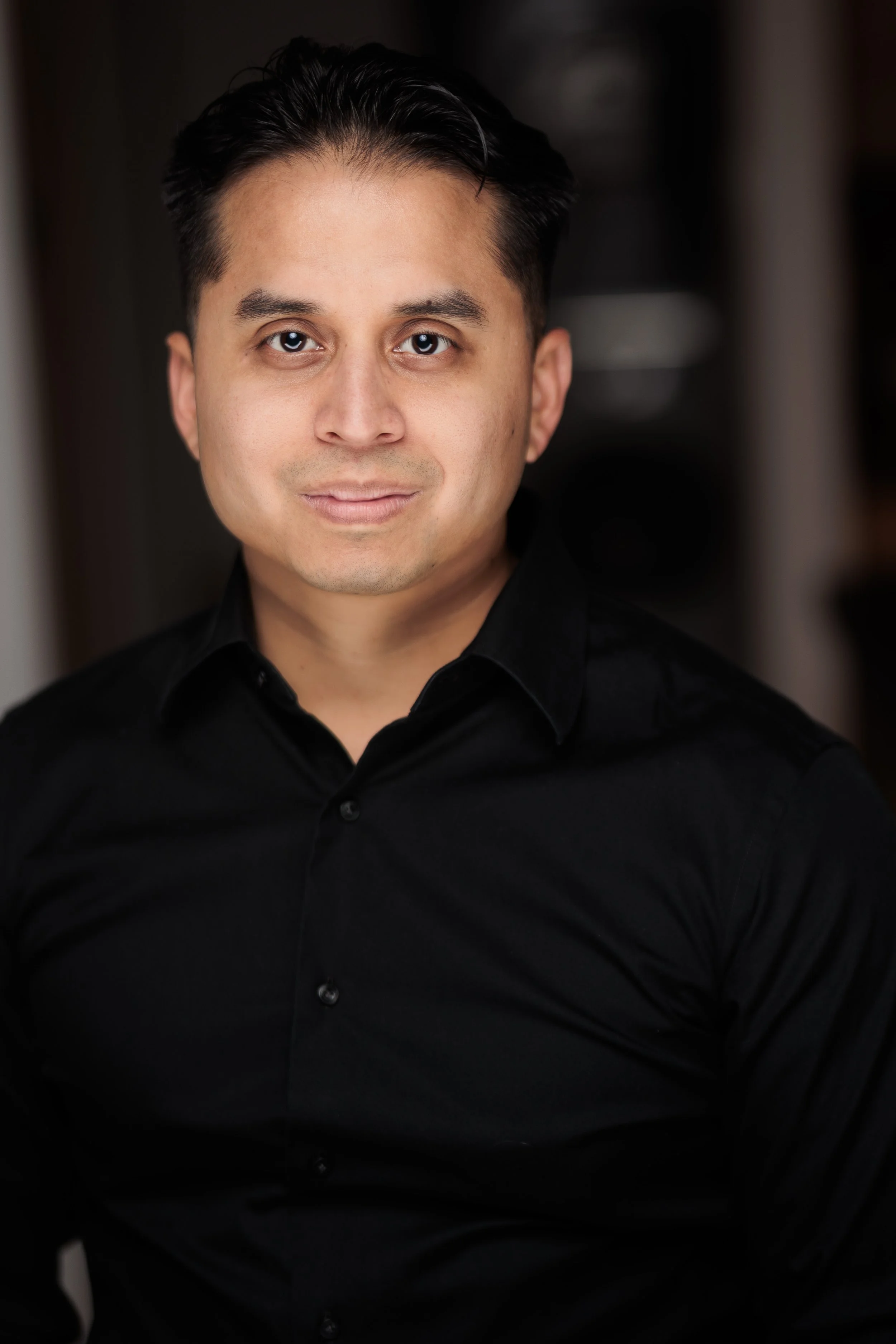 Headshot of a man with dark hair, wearing a black shirt, looking directly at the camera against a blurred dark indoor background.