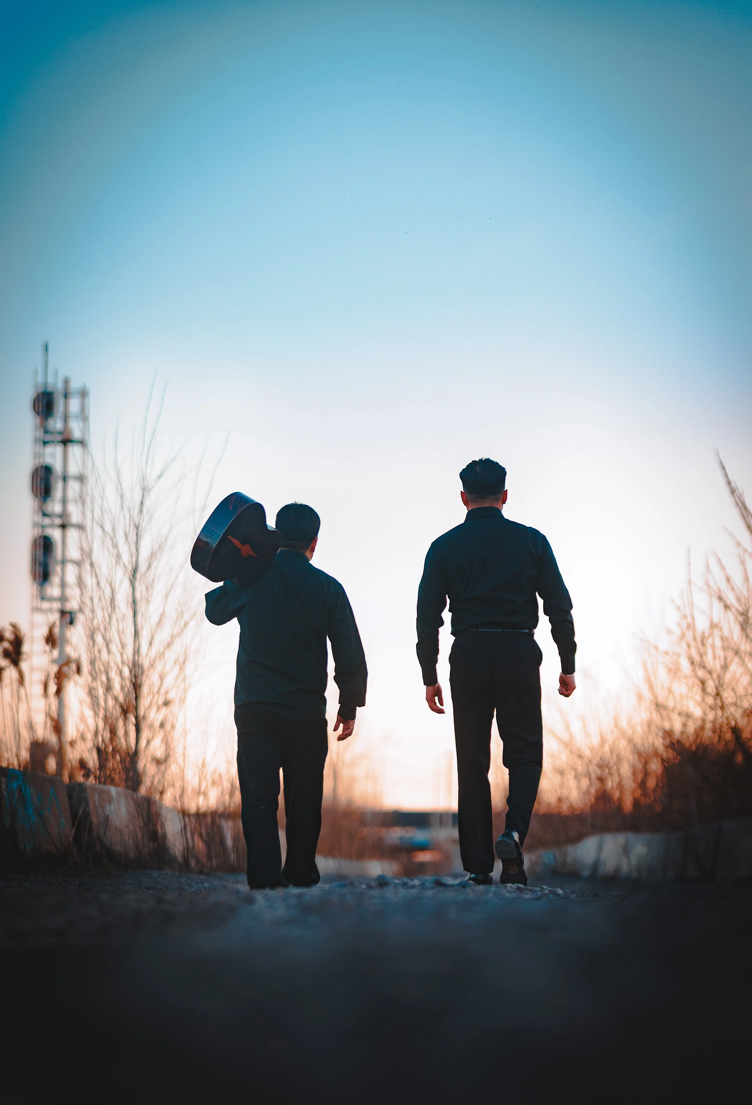 Two men walking down a dirt path during sunset, one carrying a guitar, surrounded by dry trees and power lines, with a clear blue sky overhead.