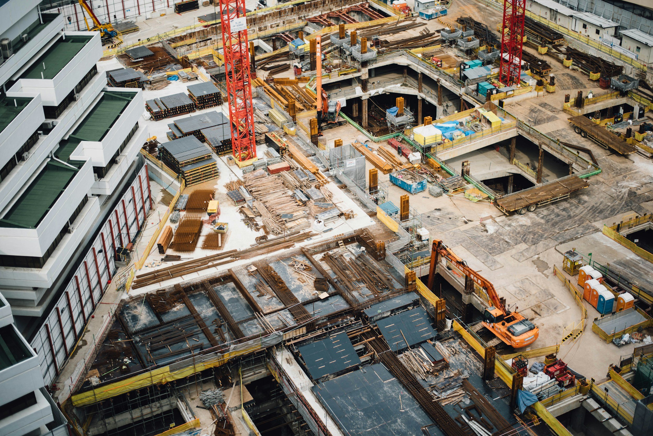 Aerial view of a large construction site with multiple cranes, building frameworks, and construction materials, adjacent to modern white buildings.
