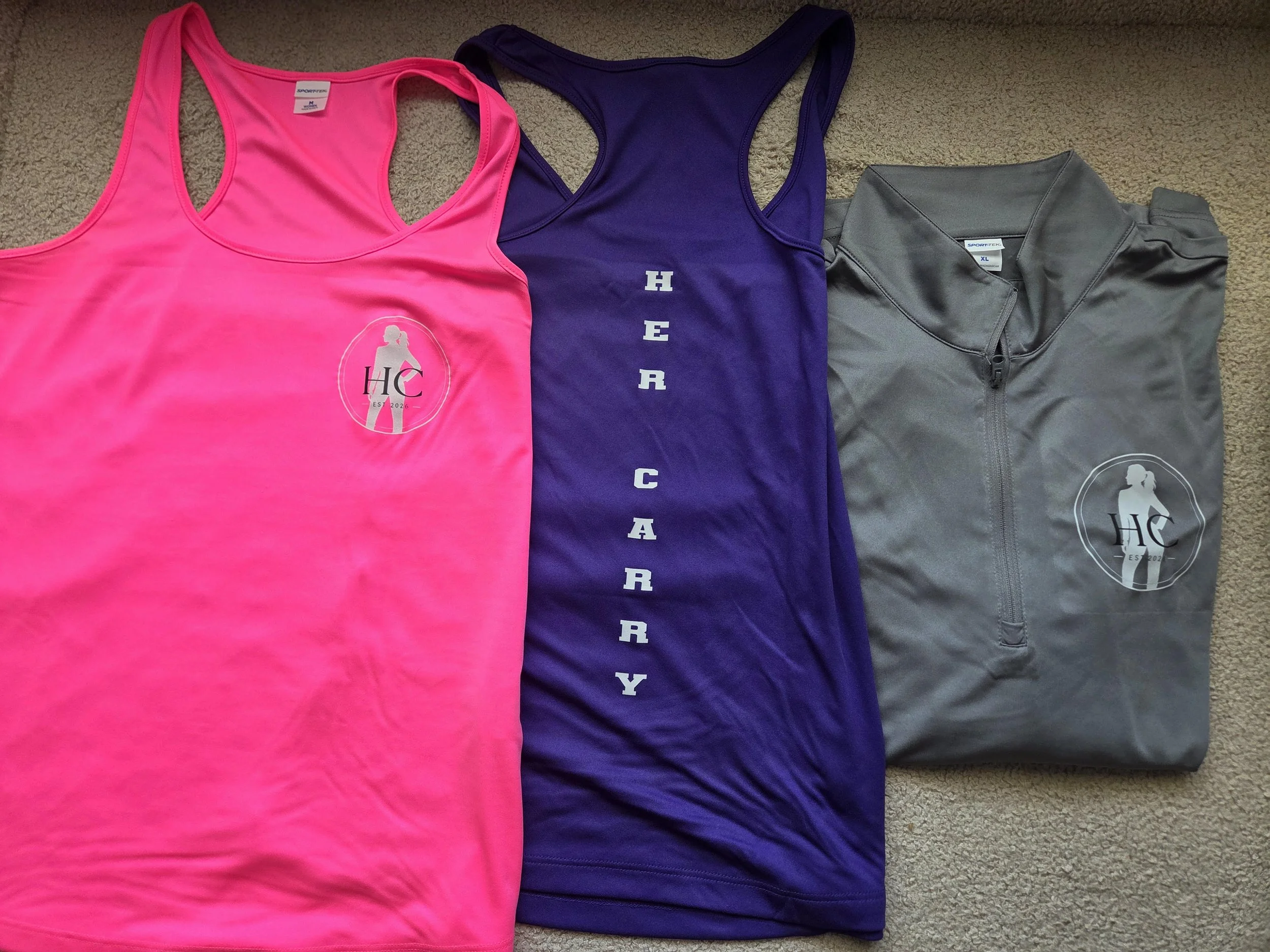 Three workout tank tops lying on a beige carpet. The first tank top is bright pink with a logo of a silhouette of a woman and the letters "HC" inside an oval, with the text "EST 2026" below. The second tank top is dark purple with vertically aligned white text spelling "HER" and striped across the front. The third tank top is gray with a zippered front and a logo of a woman with the letters "HC" and "EST 2026" underneath.