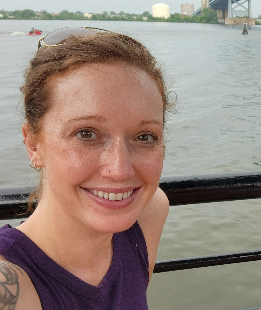 A woman with red hair smiling near a body of water, with a bridge and some industrial structures in the background.